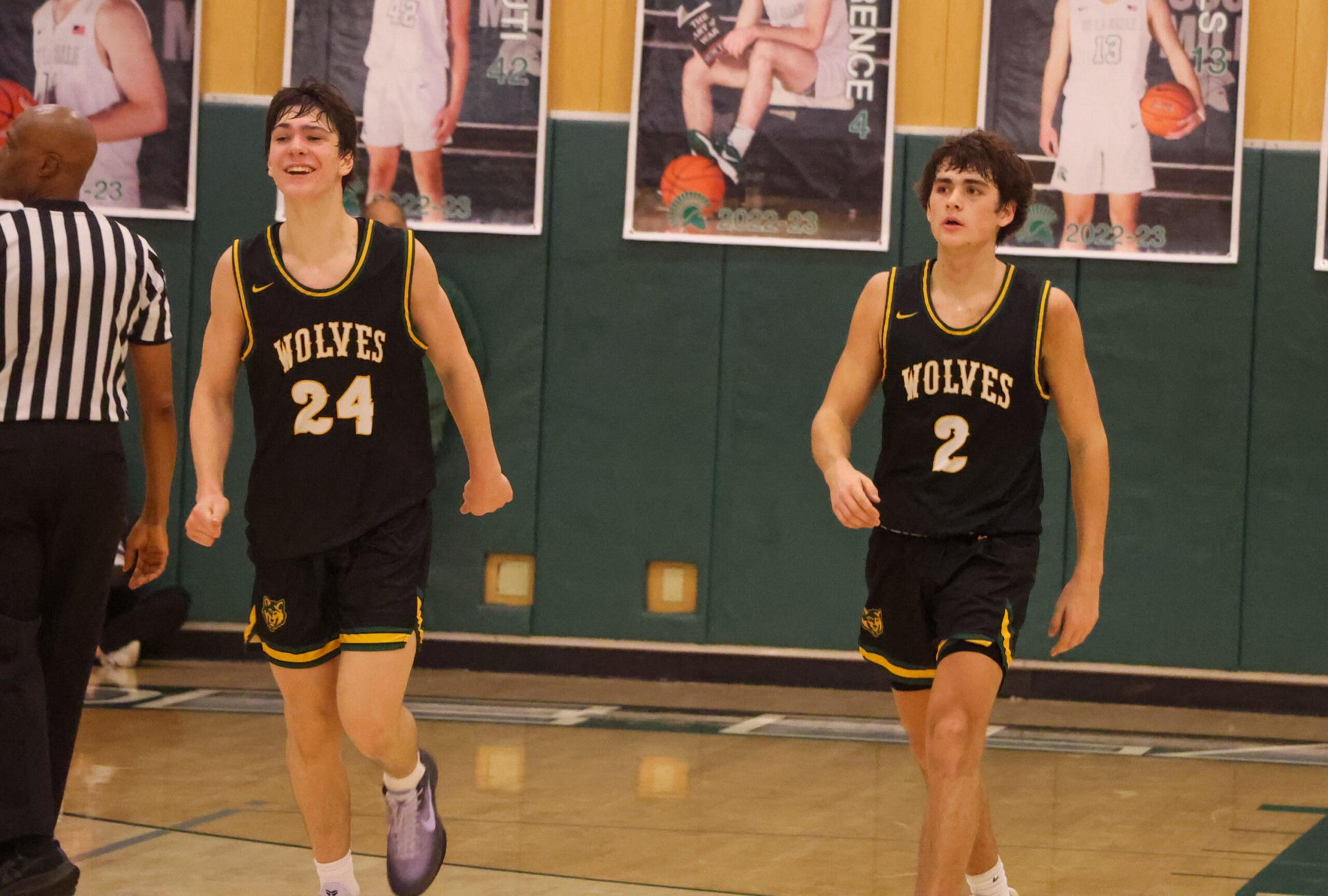 Two basketball players wearing black "Wolves" jerseys numbered 24 and 2 walk on a gym court near a referee, with sports posters visible on the wall behind them.