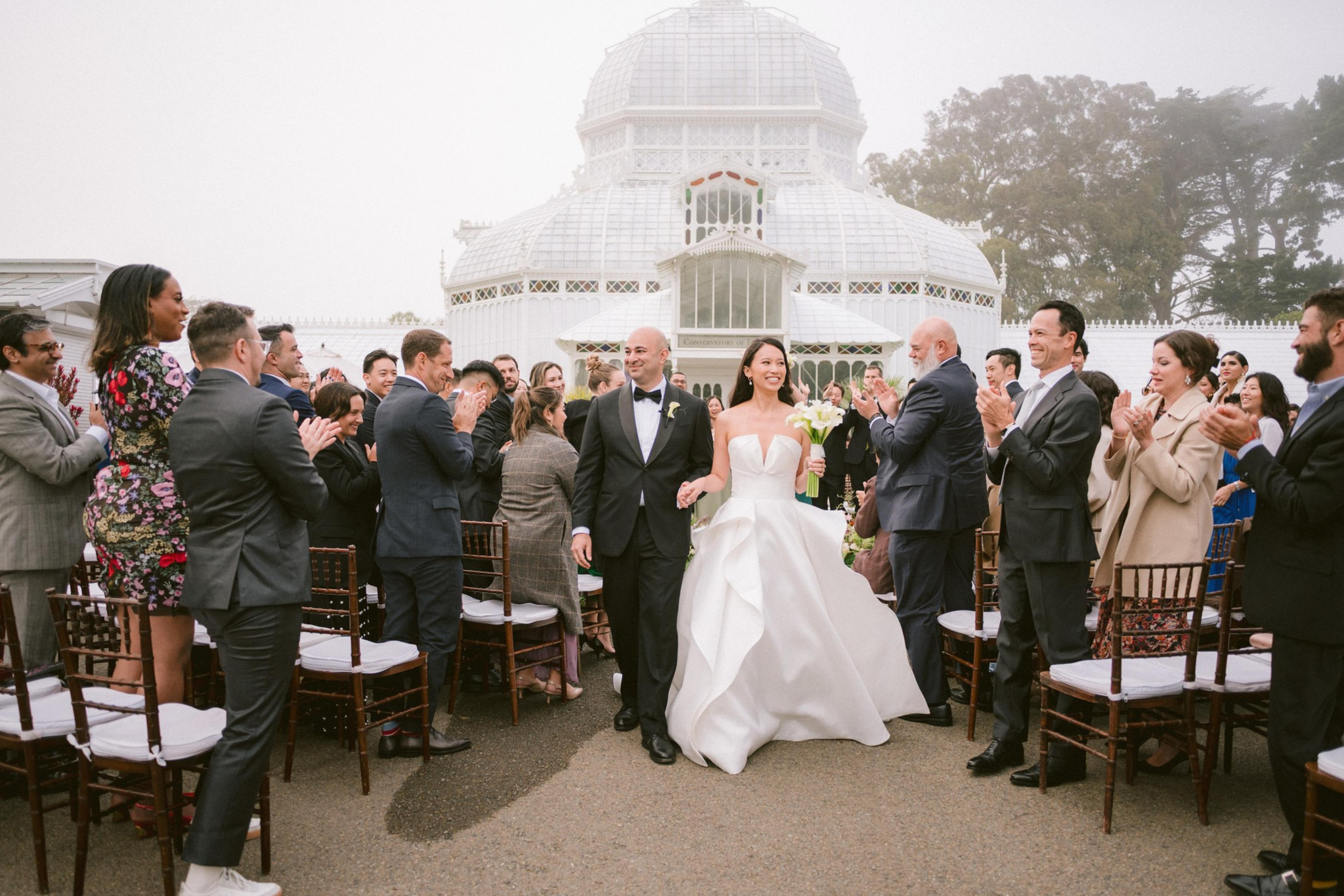 A bride in a white gown and groom in a black tuxedo walk hand-in-hand down an outdoor aisle while guests applaud.