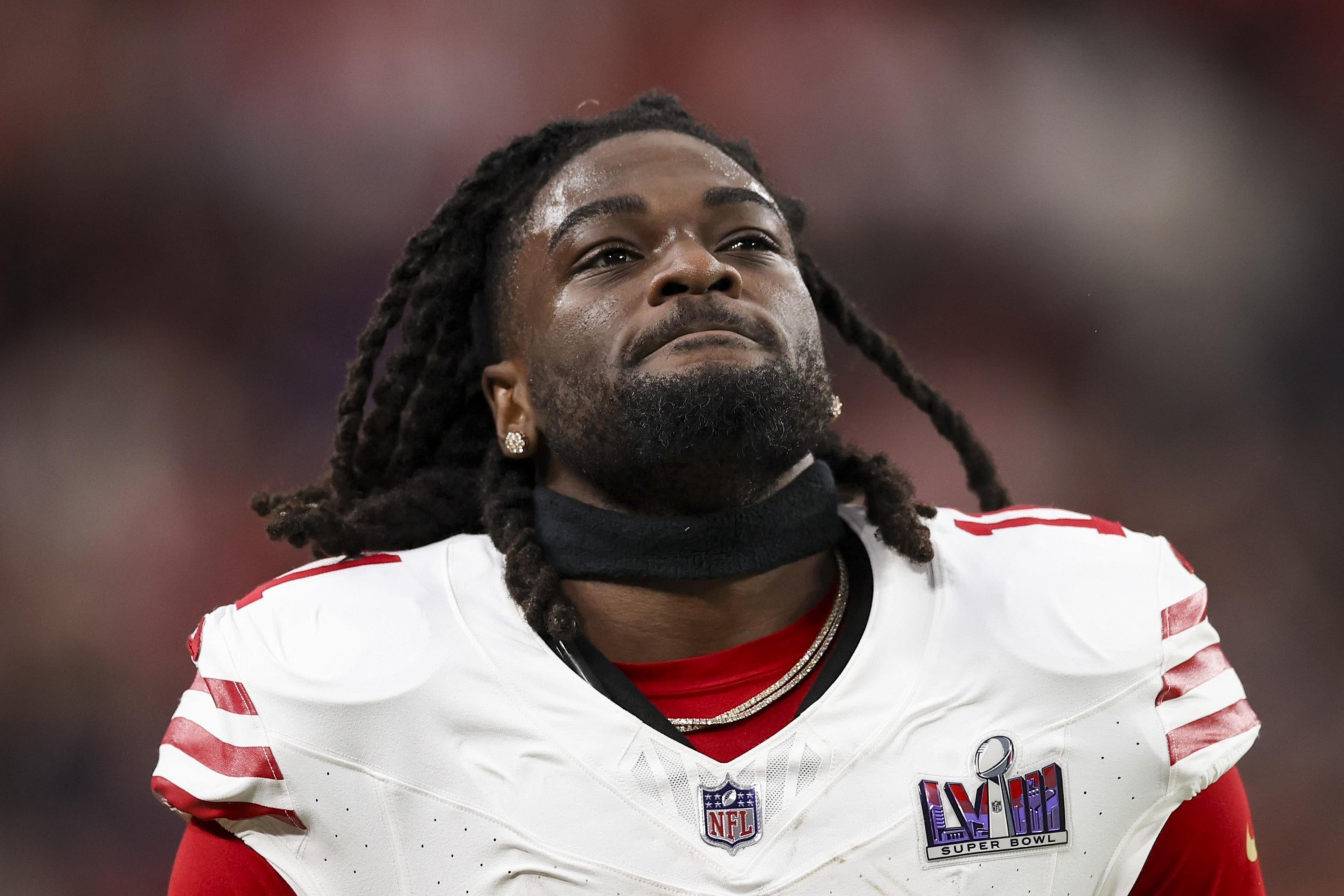 A football player wearing a white and red uniform with a Super Bowl patch gazes upwards, with long dreadlocks and earrings, on a blurred stadium background.