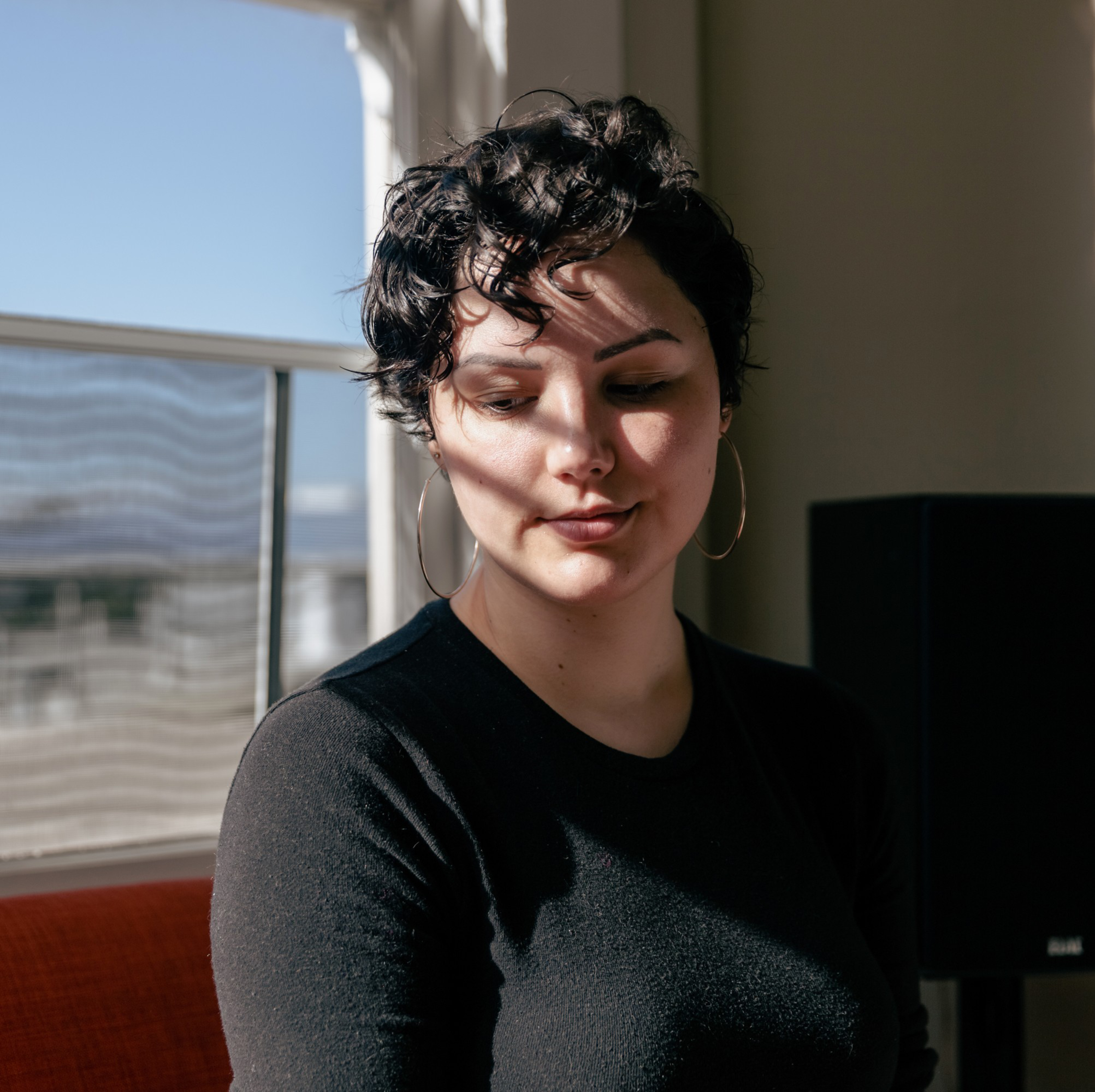 Sasha Perigo with short curly hair, large hoop earrings, and a black top is in a sunlit room.