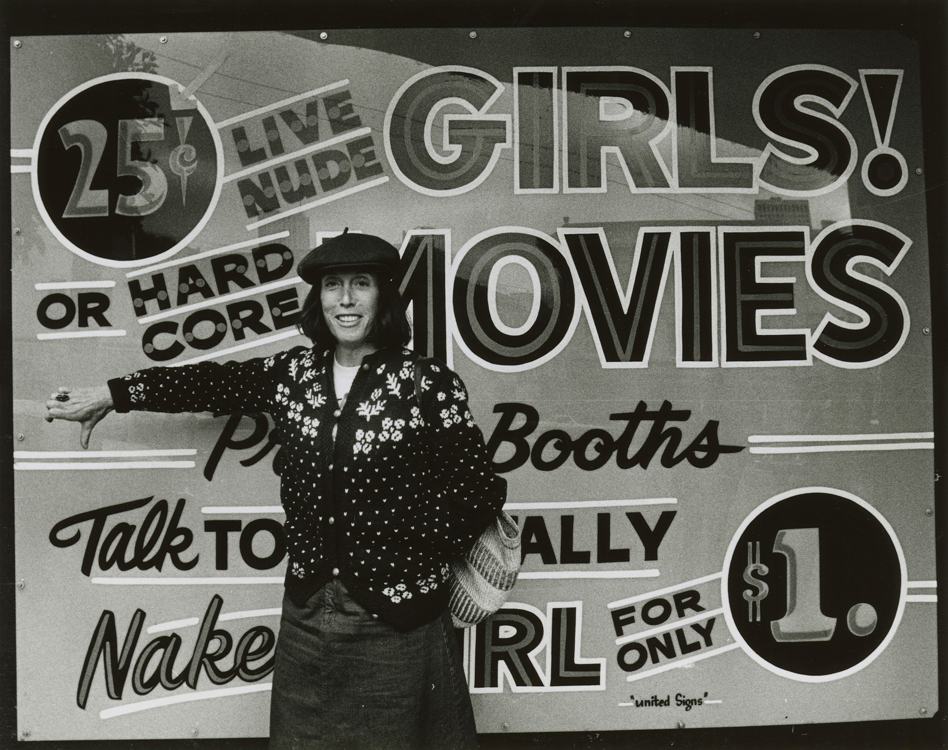 A person in a polka-dot jacket and hat stands smiling with a thumbs-down in front of a bold, colorful sign advertising "girls," "movies," and prices.