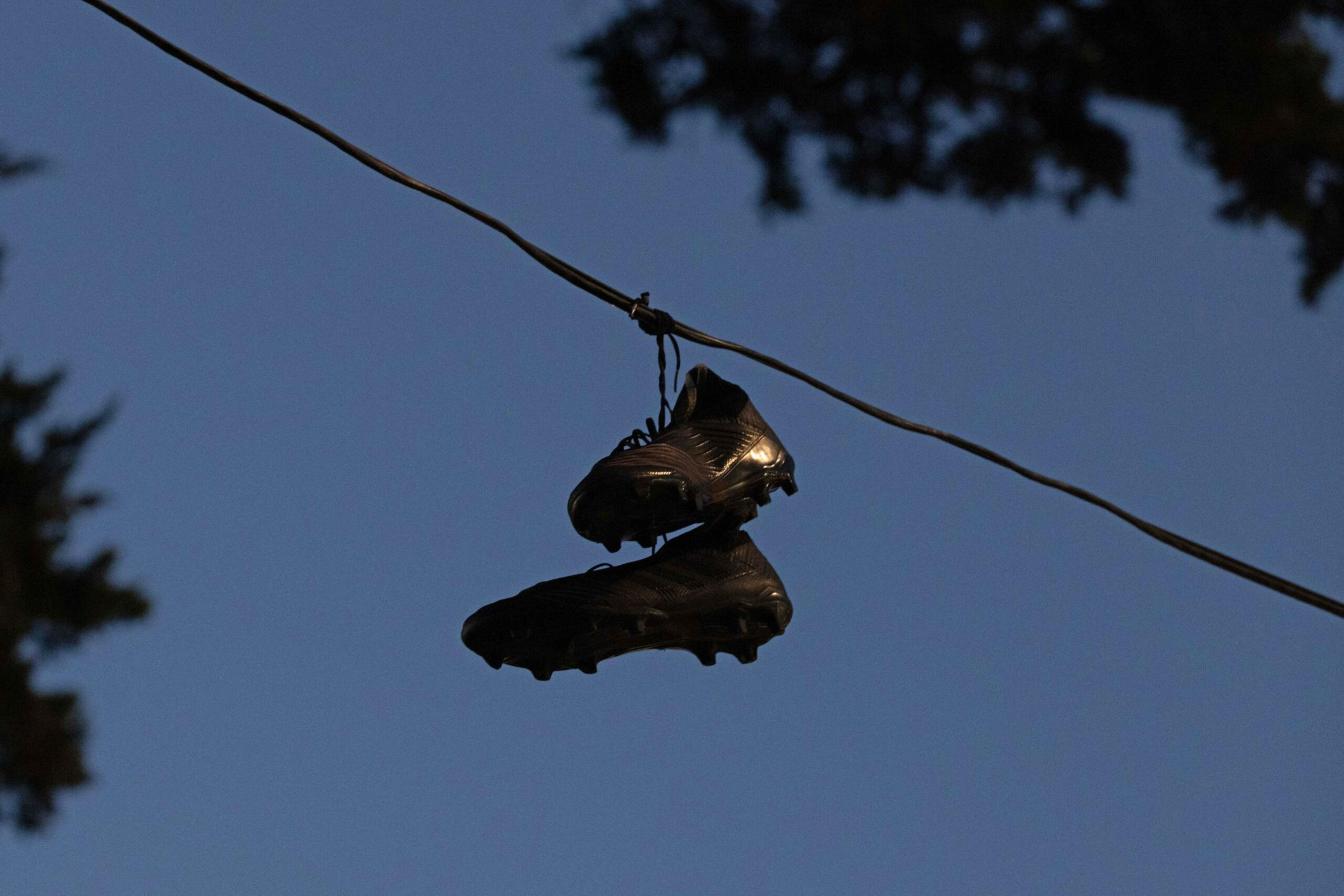 A pair of black soccer cleats is hanging by their laces over a wire, set against a clear blue sky, with partial silhouettes of trees in the background.