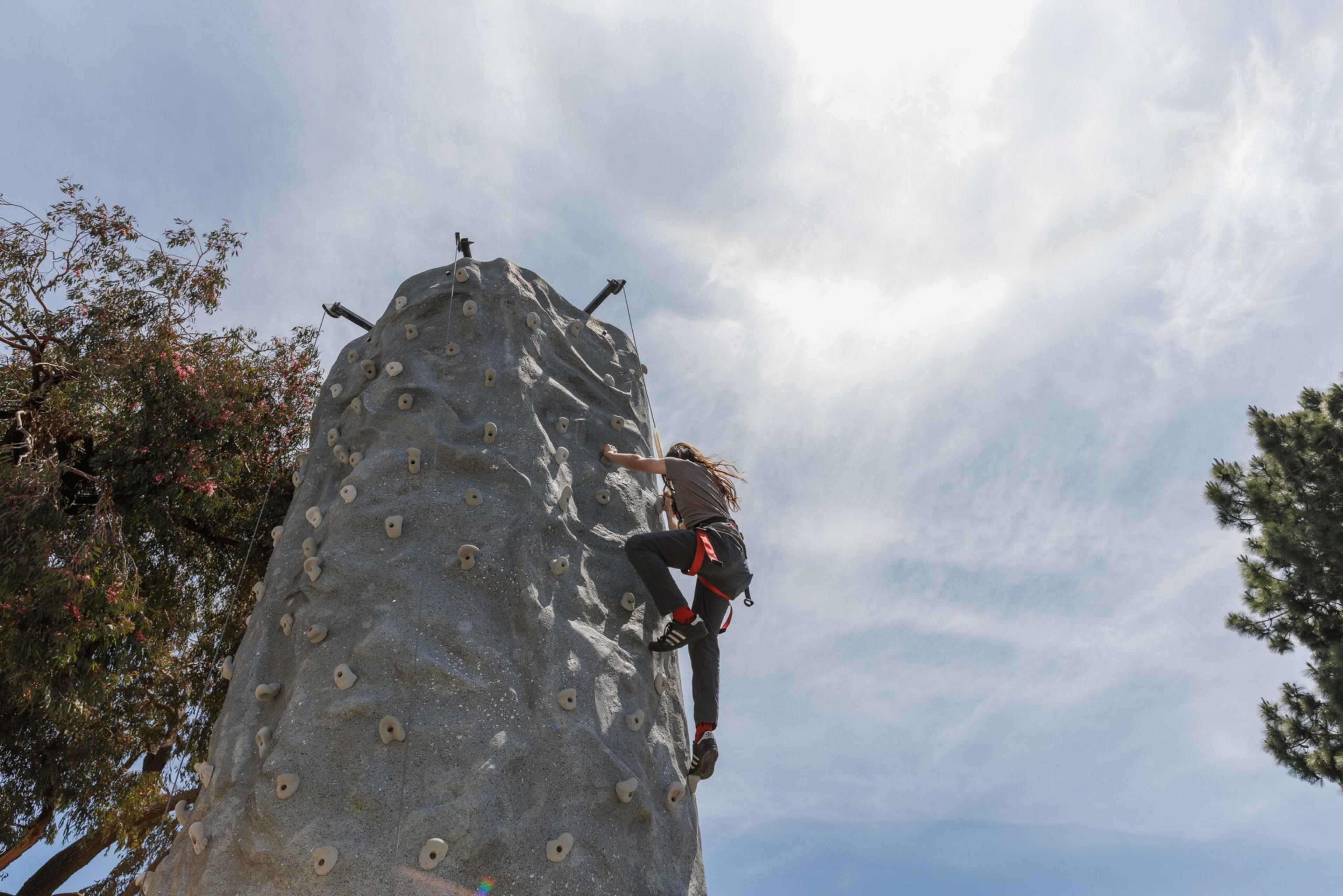 A person is scaling a climbing wall against a backdrop of clouds and trees.
