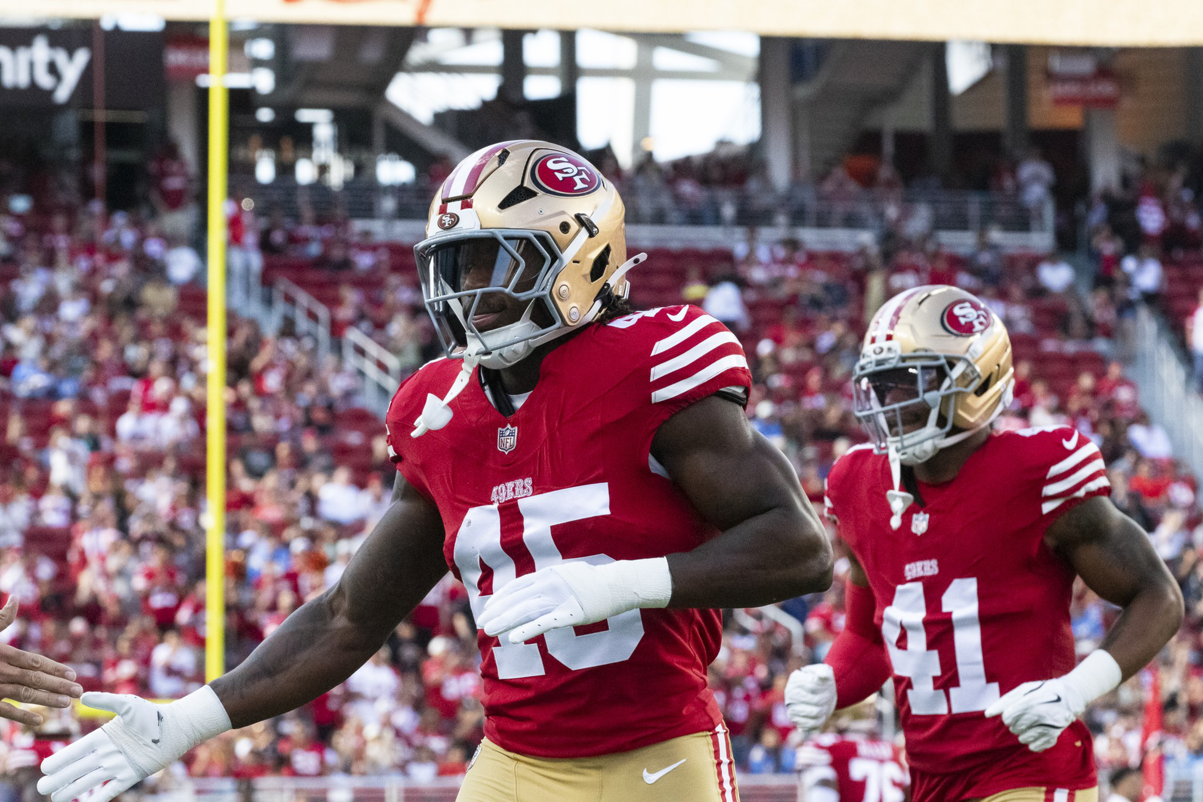 Two football players in red and gold San Francisco 49ers uniforms celebrate on the field with a crowd in the background.