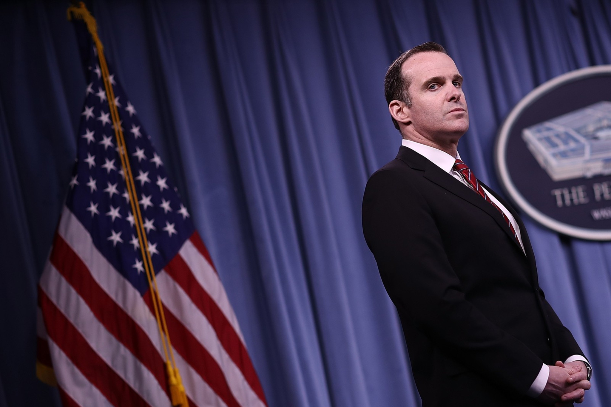 A man in a black suit stands with folded hands against a blue curtain backdrop, near an American flag and a Pentagon emblem.