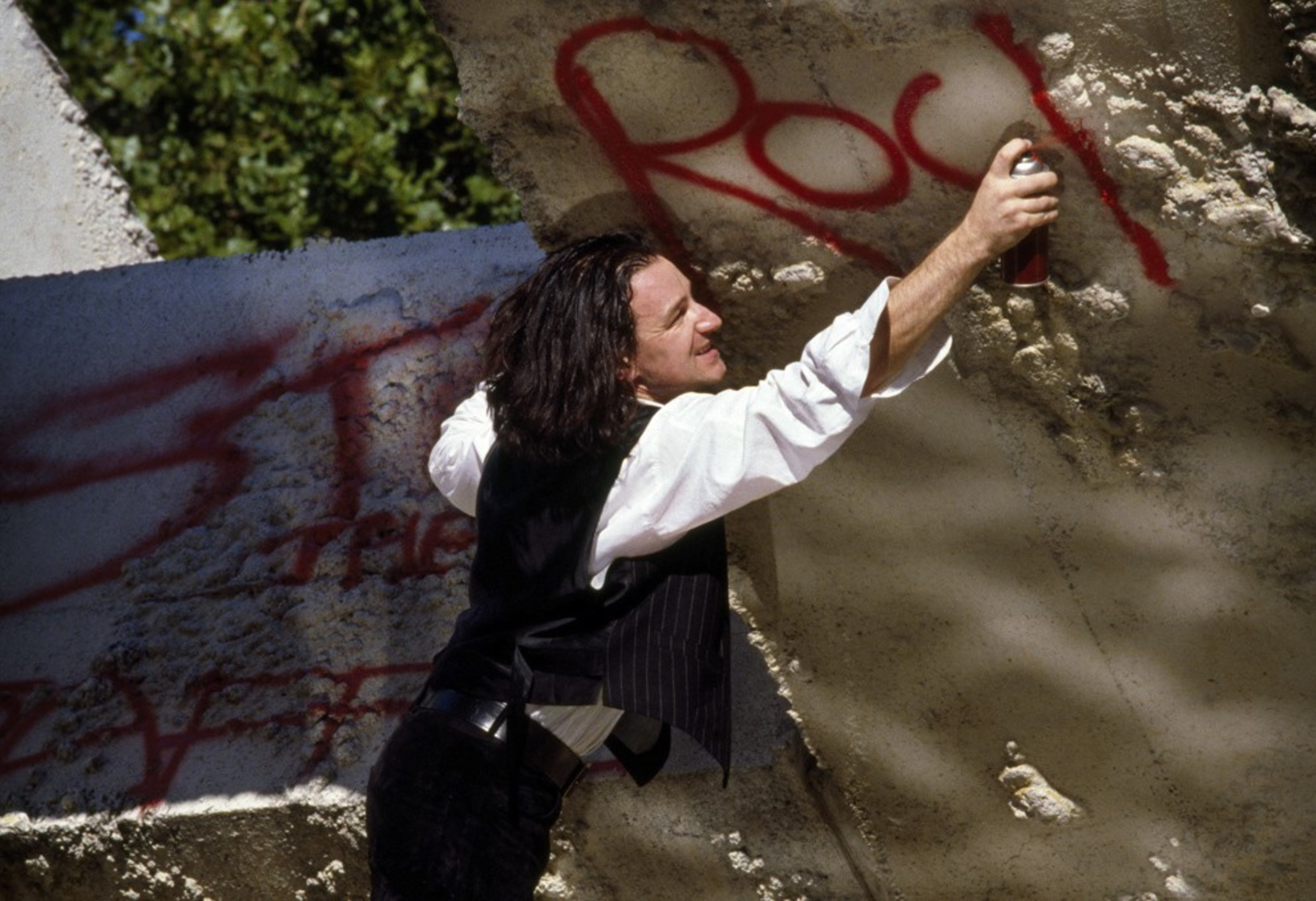 A person with long hair spray-paints the word &quot;Rock&quot; in red on a textured concrete wall. They wear a white shirt and black vest. Trees are visible in the background.