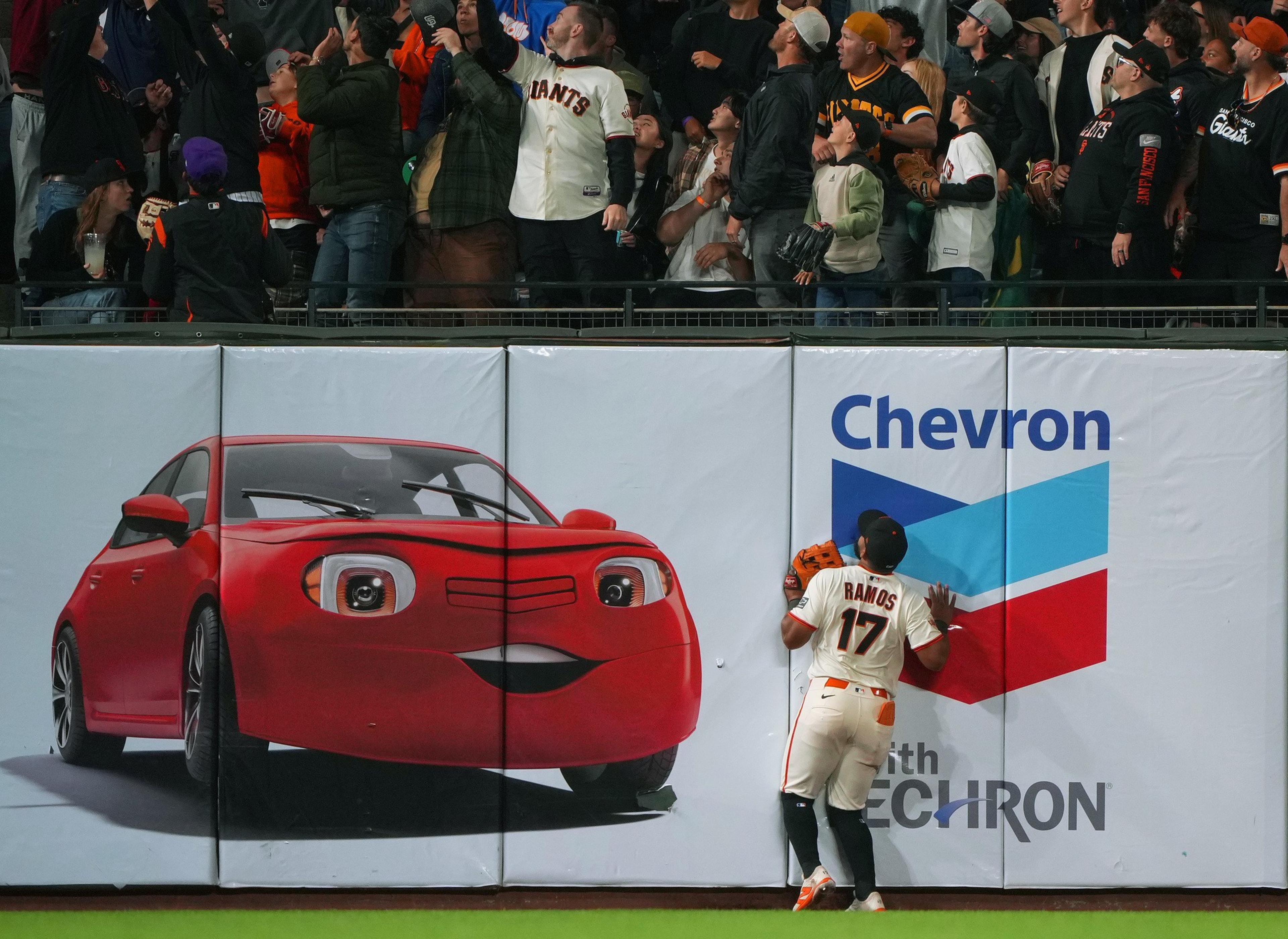 A baseball player labeled "Ramos 17" leans against an outfield wall near a large advertisement of a red cartoon car with expressive eyes and a smiling mouth, as fans watch.