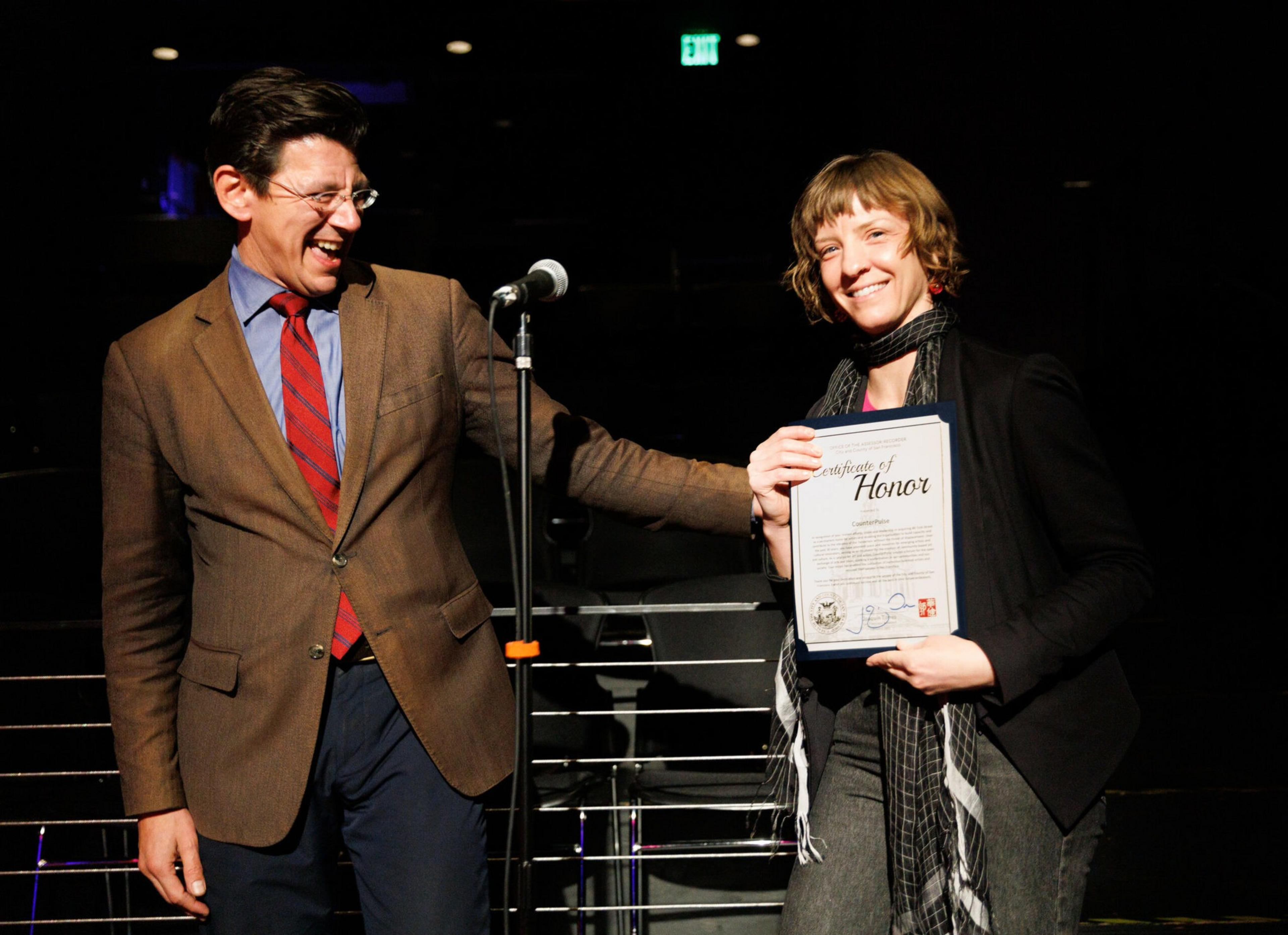 A man in a brown suit laughs while a woman in a black jacket smiles, holding a "Certificate of Honor" in her hands on a dimly lit stage.