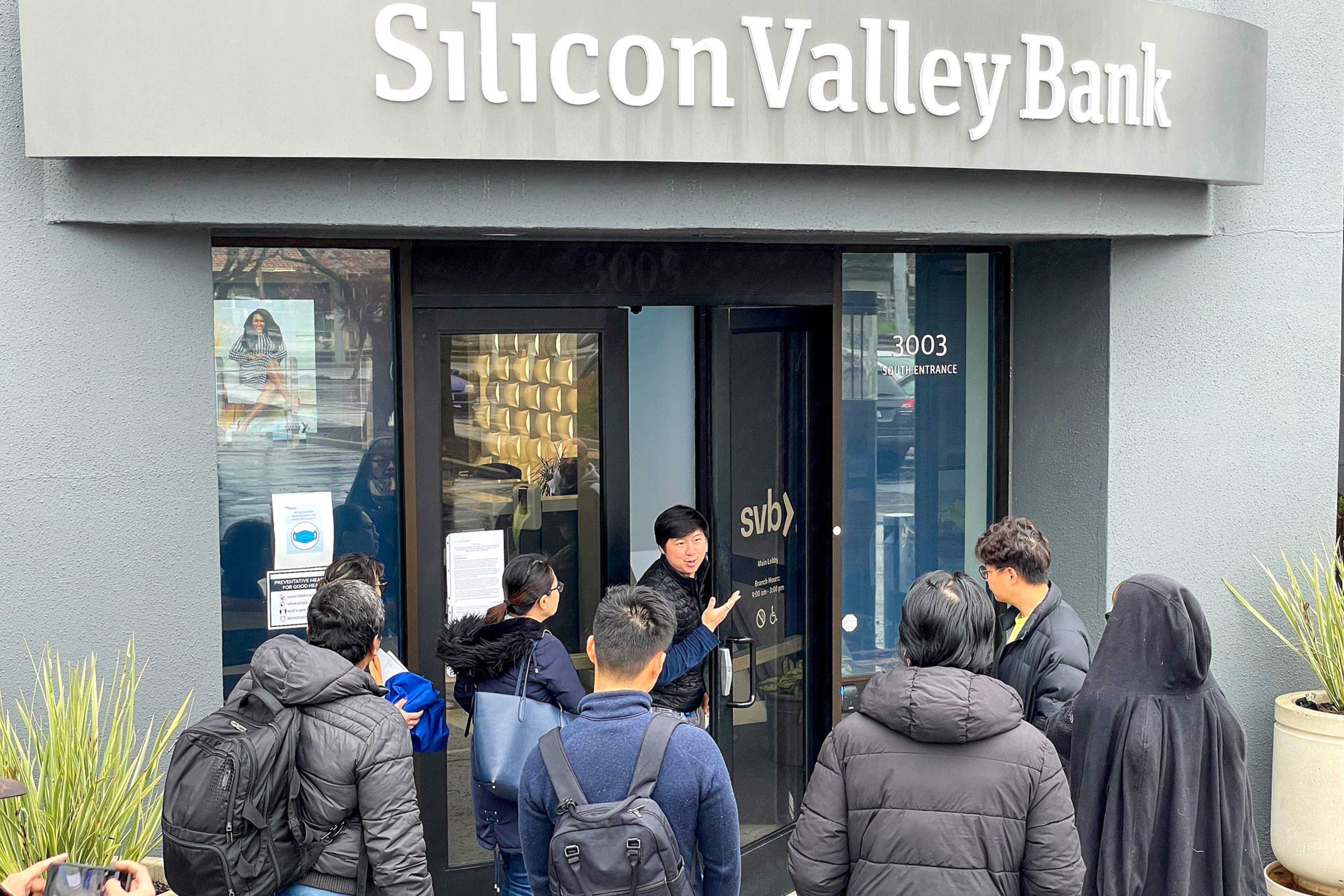 A group of people stand outside the Silicon Valley Bank entrance, where a person gestures as if speaking to them.