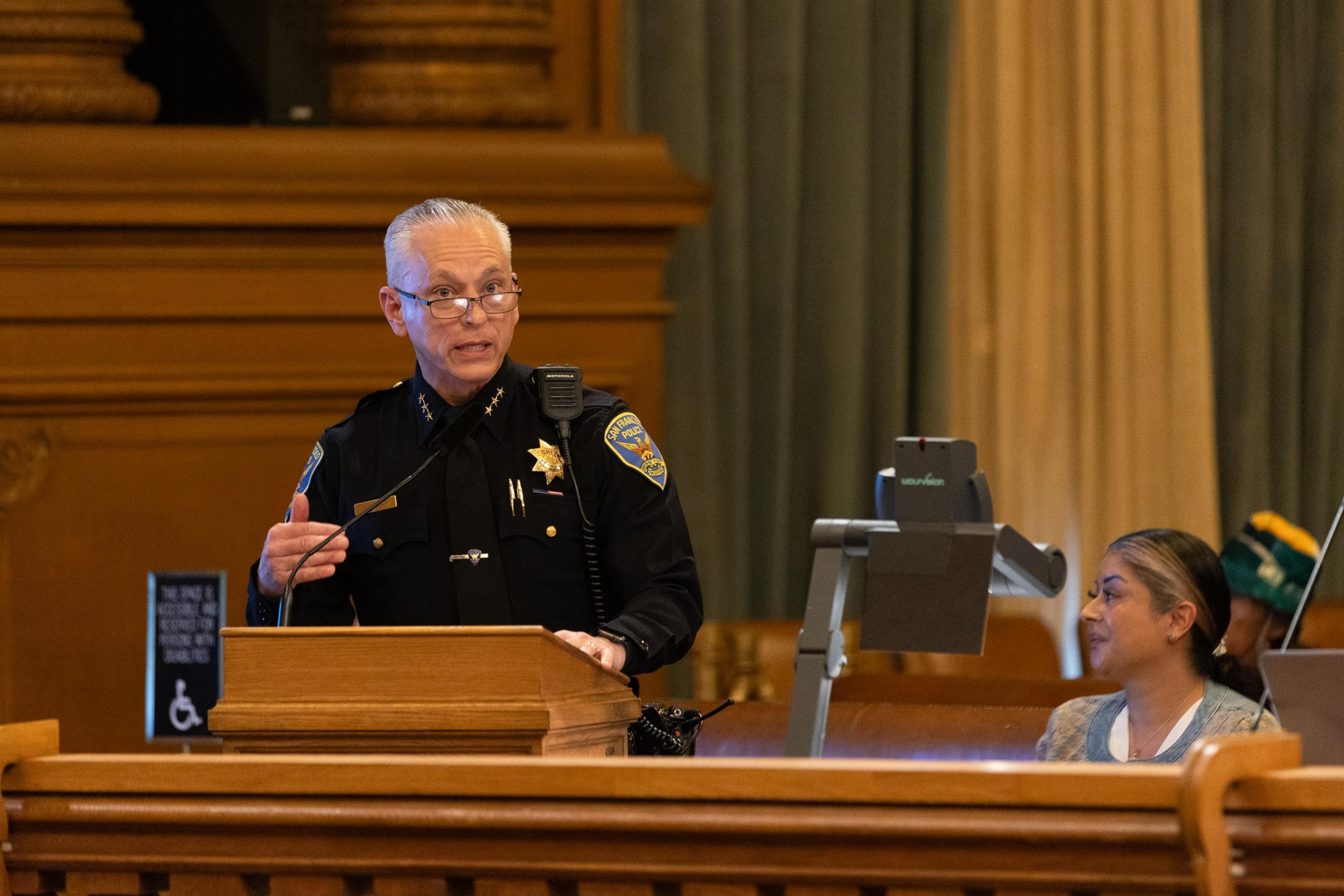 A police officer speaks at a podium in a formal setting, wearing a uniform with badges. A woman sits at a desk with a document camera next to her.