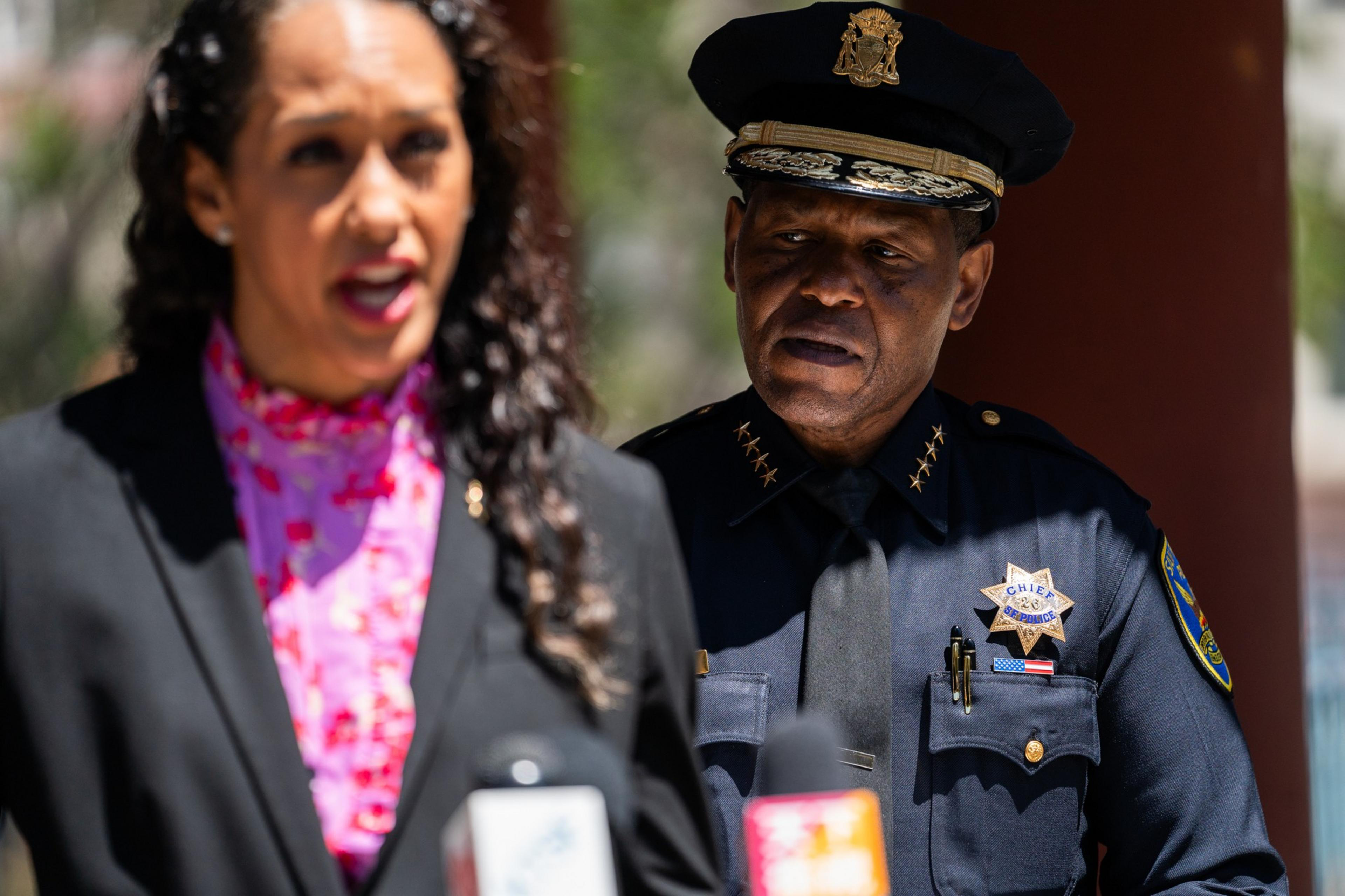 A police chief in uniform listens while a woman in a black blazer and pink blouse speaks at a microphone during an outdoor event.