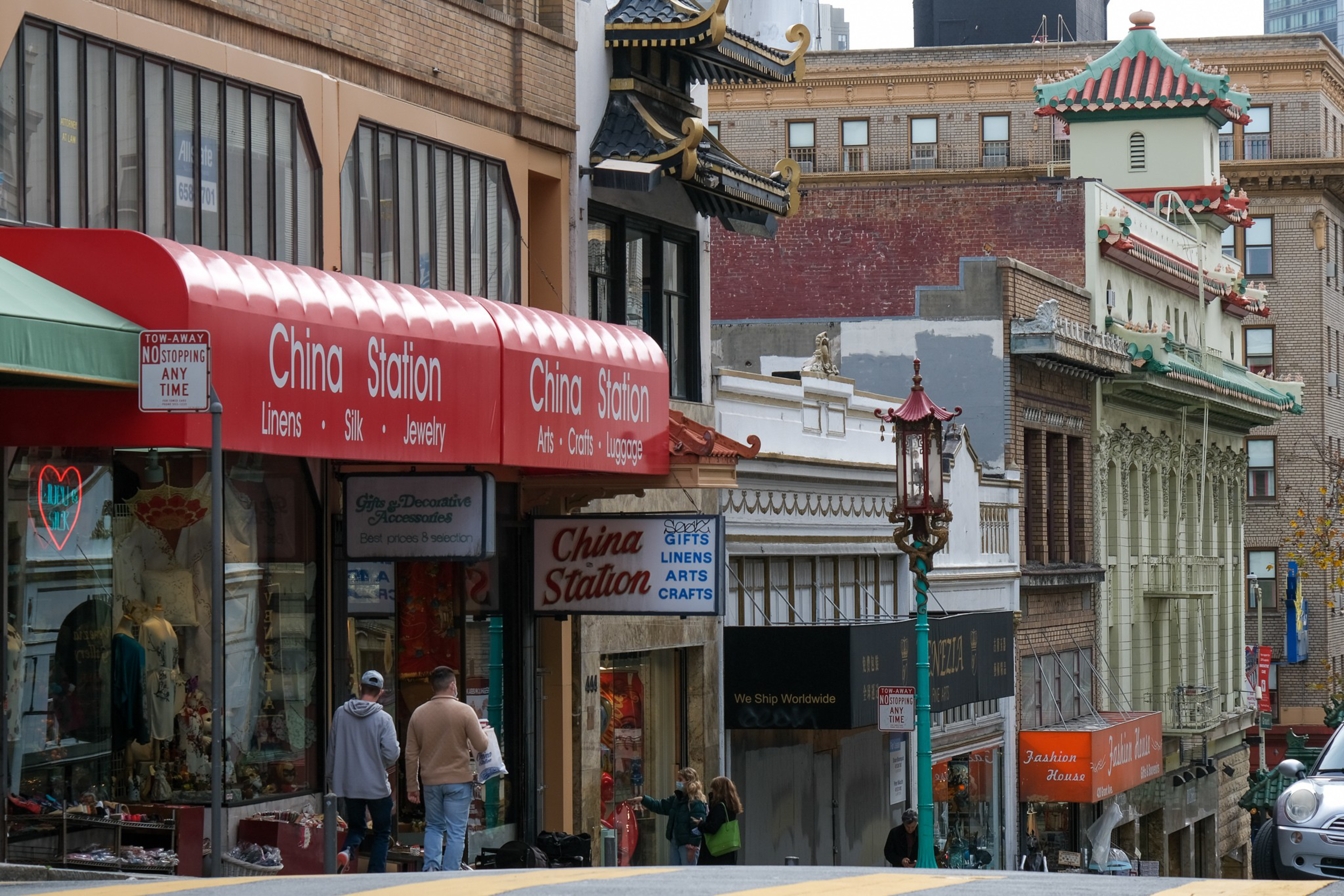 A bustling street with red awnings labeled "China Station," showcasing shops selling linens, silk, jewelry, arts, crafts, and luggage, with ornate East Asian architectural details.