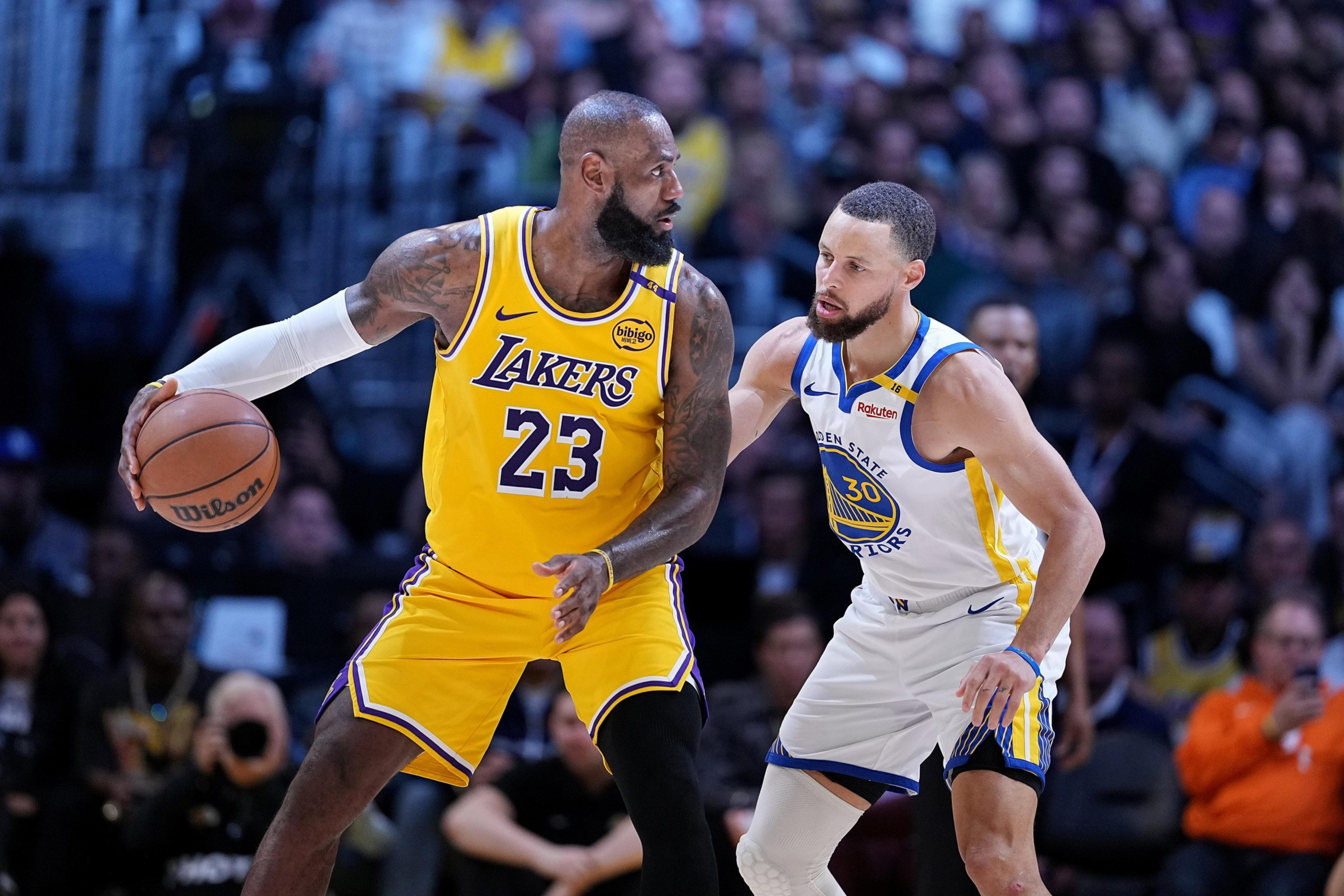 A basketball player in a yellow Lakers jersey holds the ball while being closely guarded by a player in a white Golden State Warriors jersey.