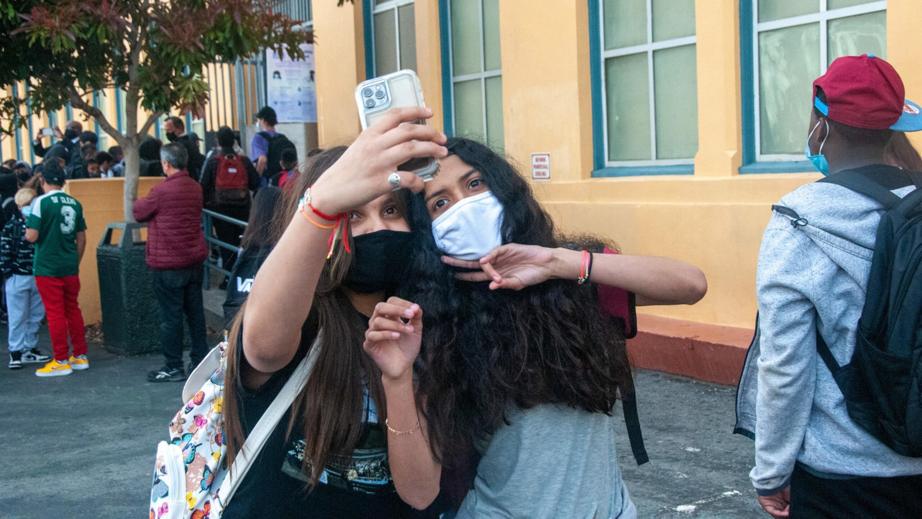 Two masked girls take a selfie outside a building, with one girl making a peace sign; a group of people stands in line in the background.