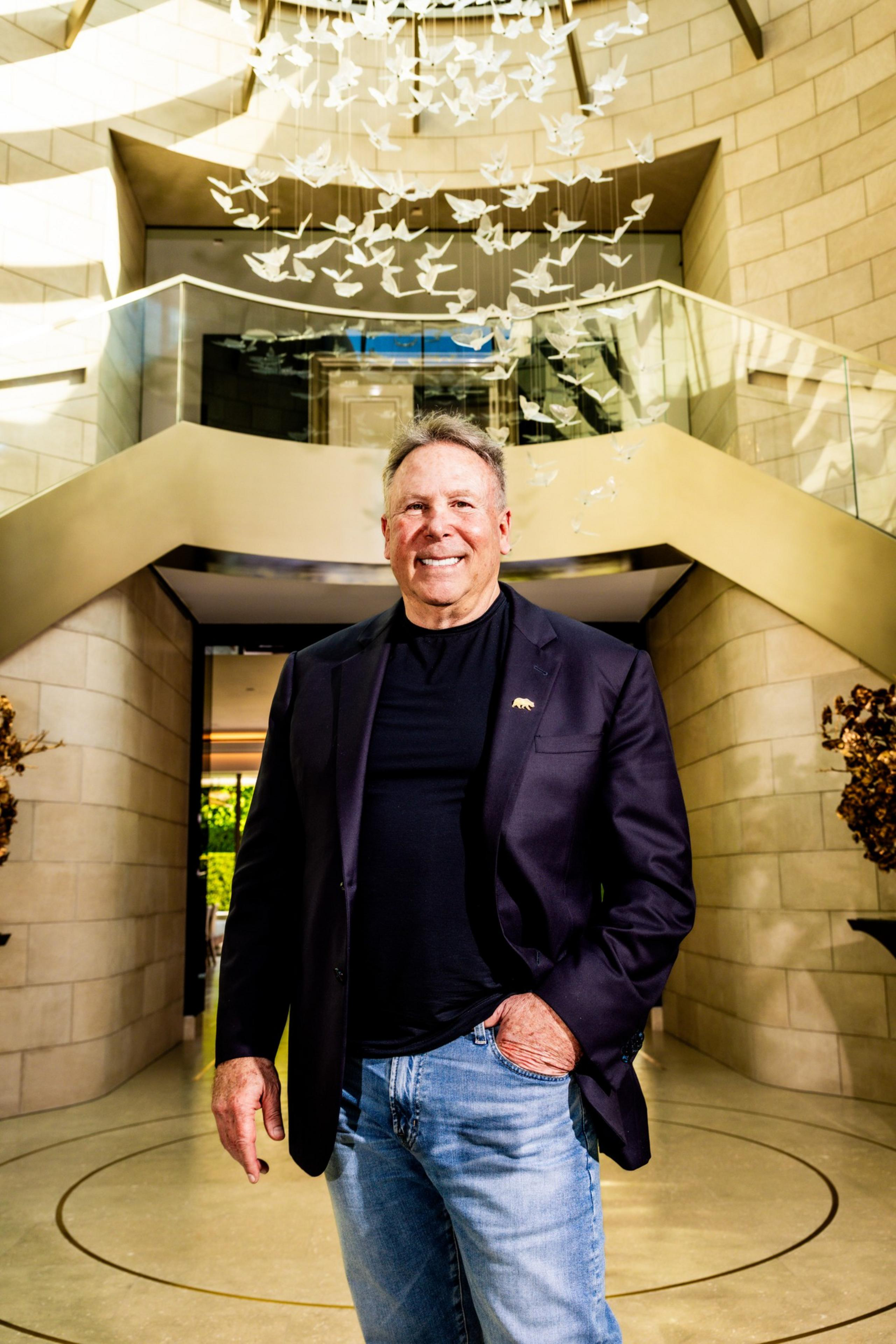 A smiling man in a black blazer and jeans stands indoors under a ceiling display of hanging white bird sculptures.