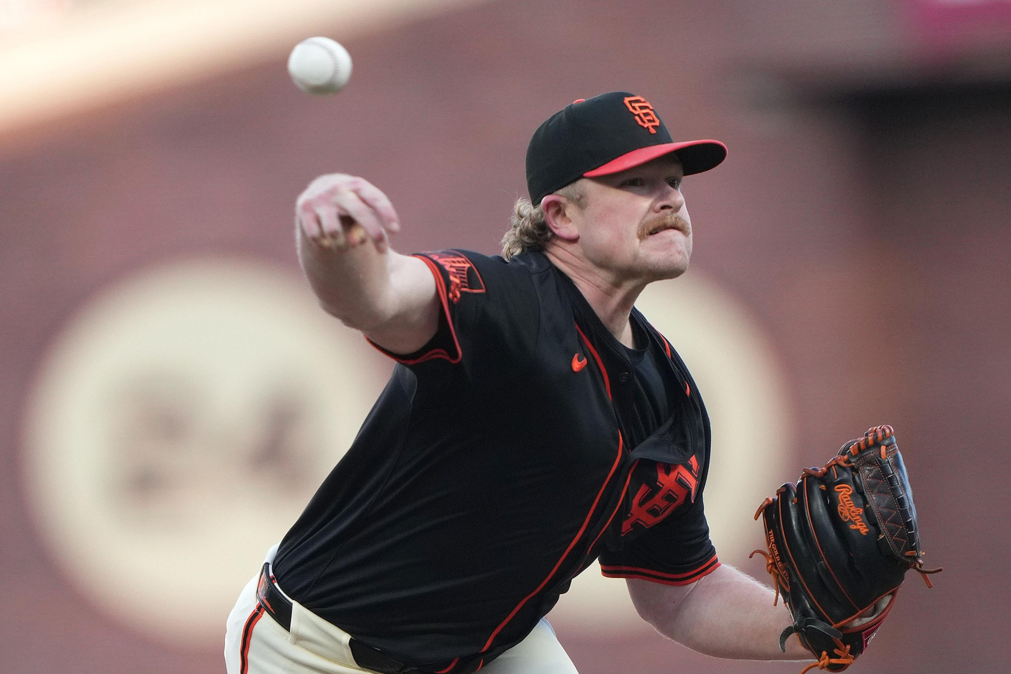 A baseball pitcher in a black and orange San Francisco Giants uniform throws a pitch with intensity, his glove hand extended forward.