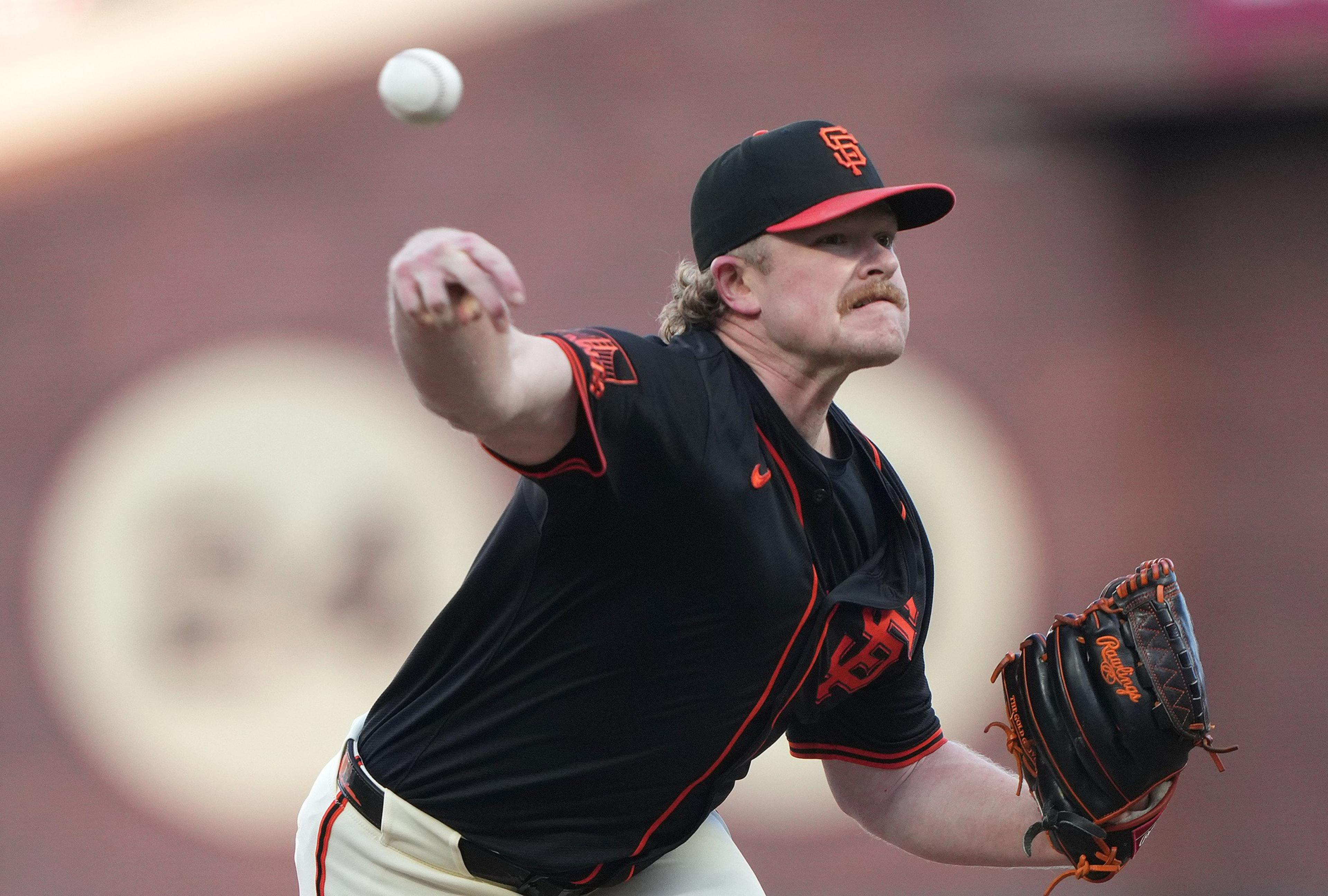 A baseball pitcher in a black and orange San Francisco Giants uniform throws a pitch with intensity, his glove hand extended forward.