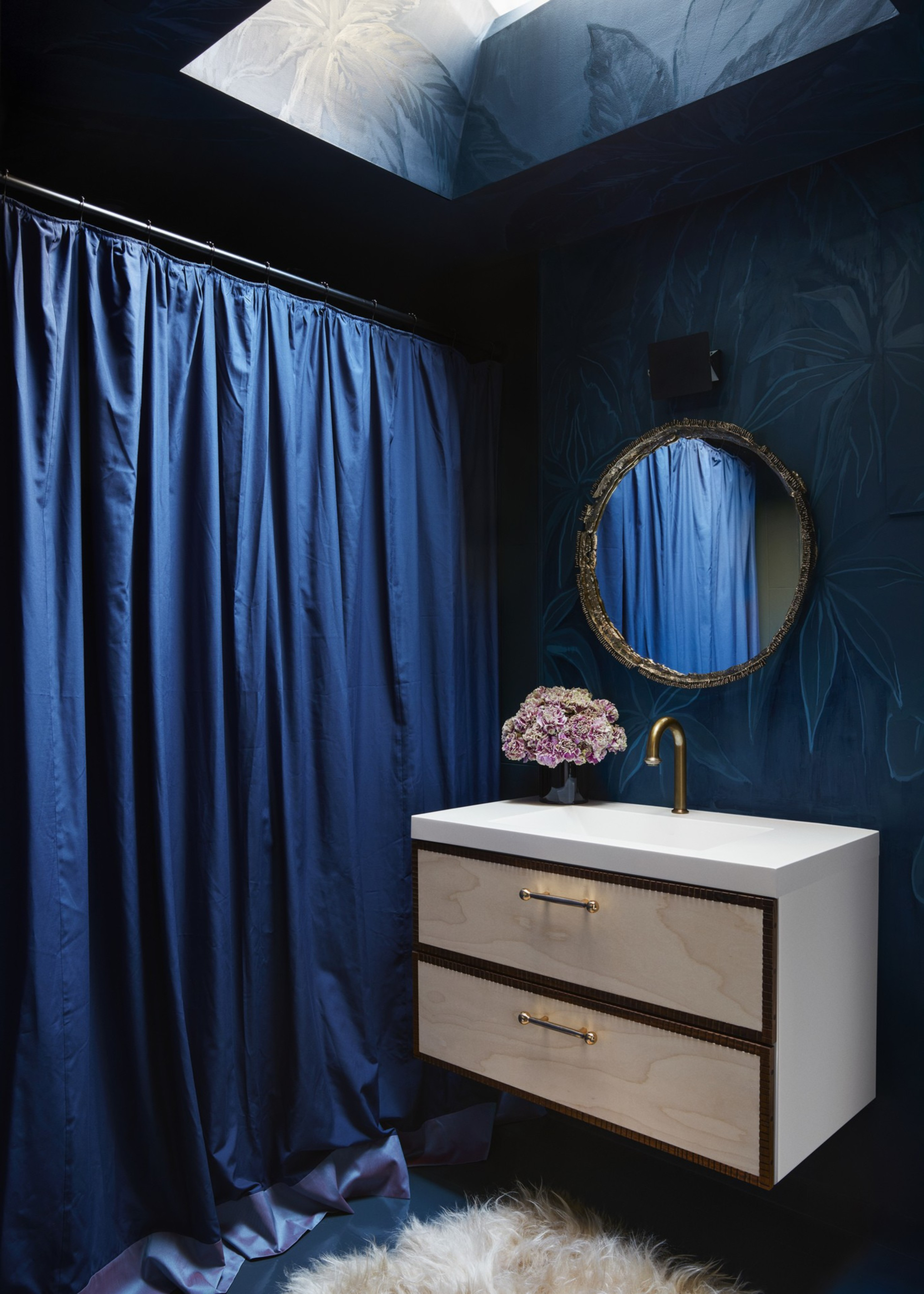 A deep blue bathroom with a shiny curtain, white sink cabinet with gold handles, a round ornate mirror, a gold faucet, a flower vase, and a fluffy white rug.