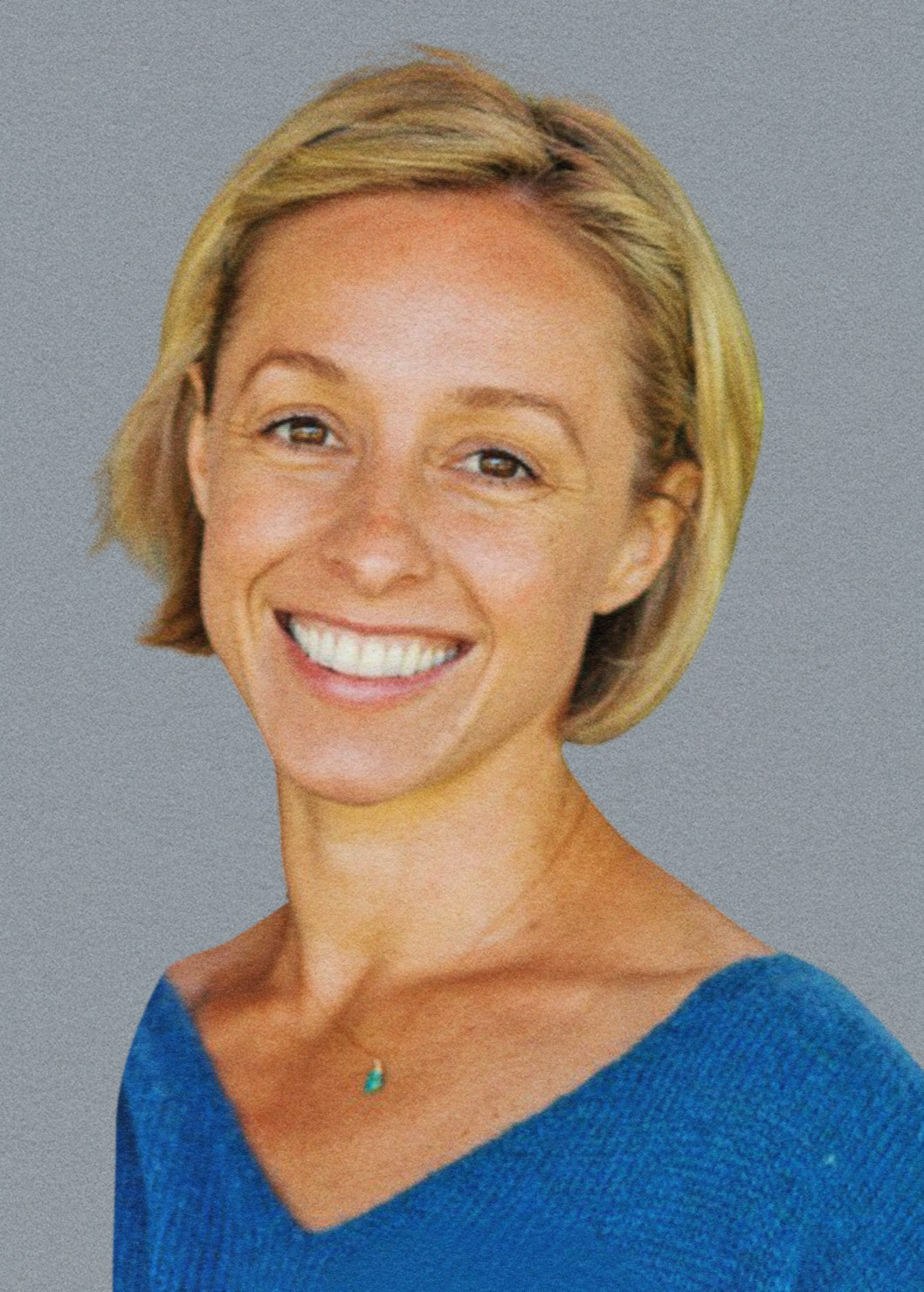 A smiling woman with short blonde hair wears a blue top and a small turquoise pendant necklace against a plain gray background.