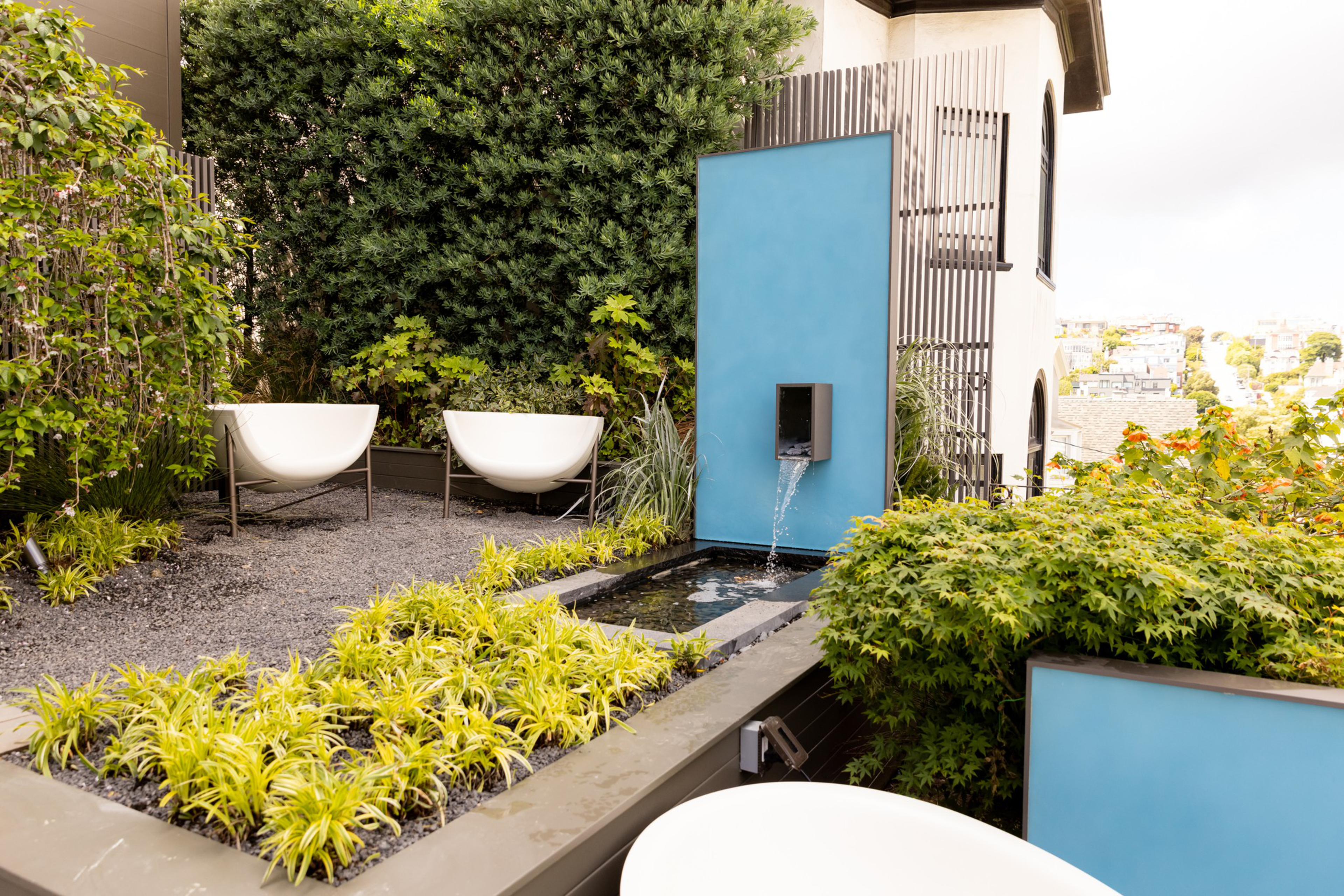 Two modern white chairs face a blue wall fountain with water flowing into a rectangular pond, surrounded by green plants in an urban garden setting.