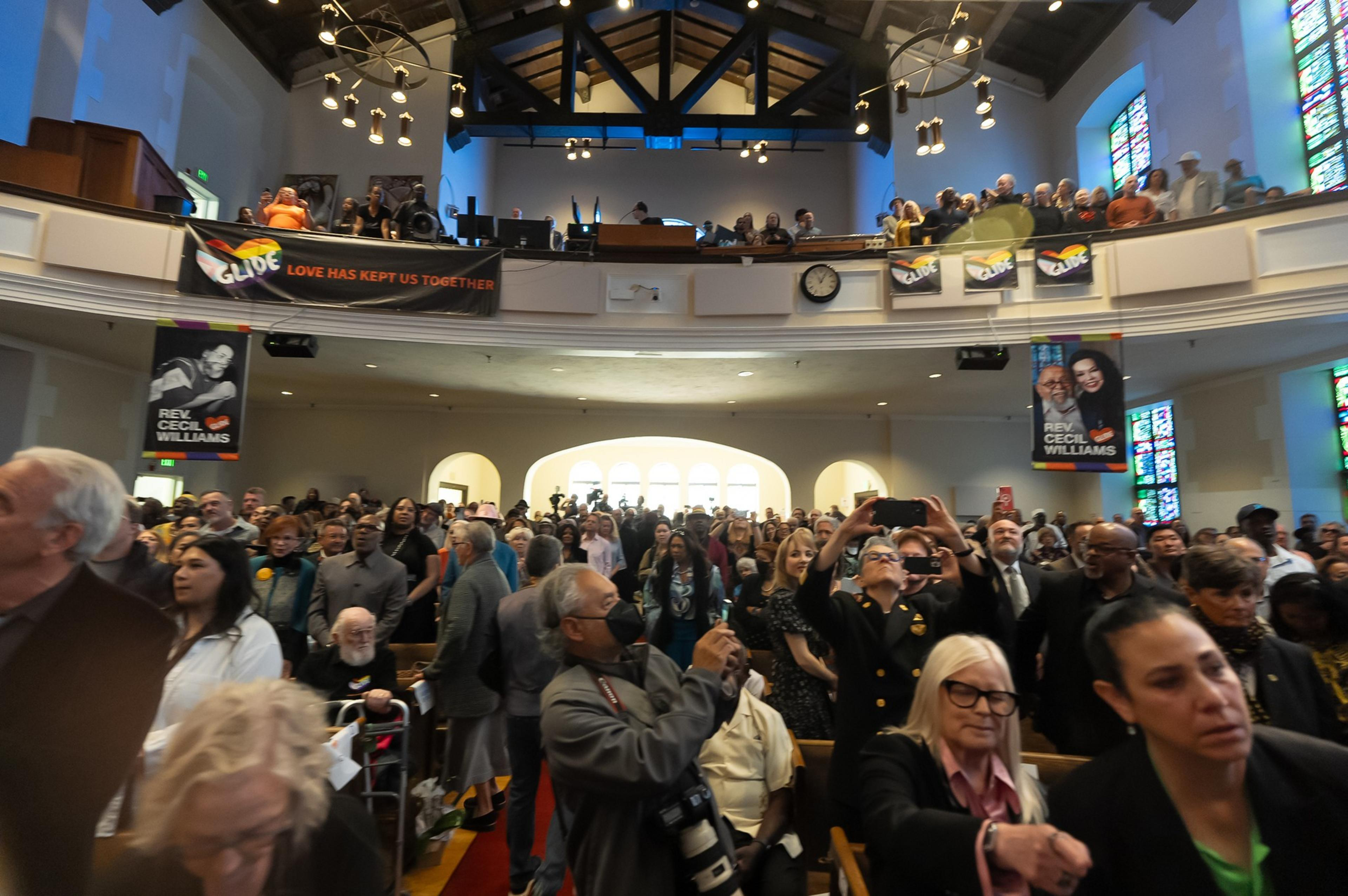 A crowded church interior with people standing, some taking photos, and banners hanging from a balcony.