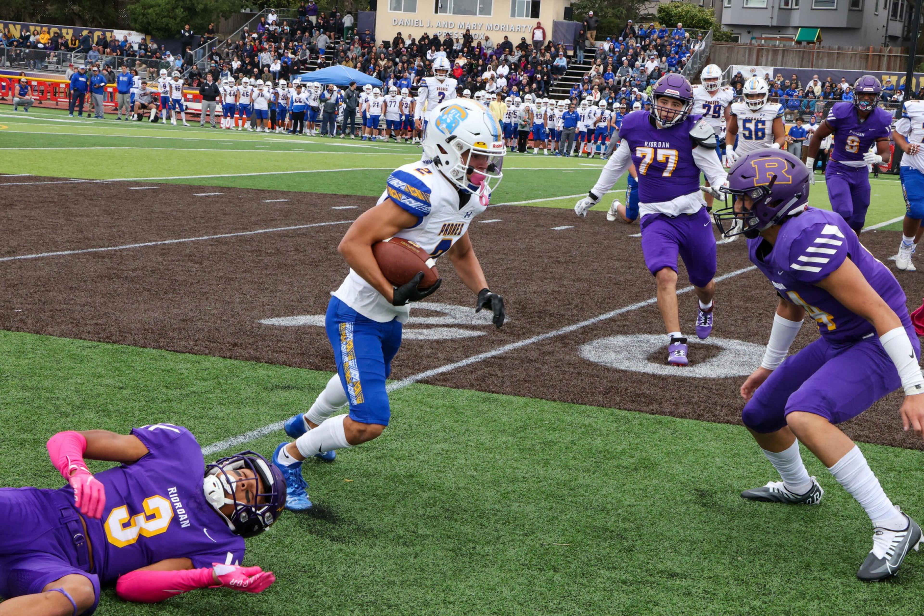 A football player in white and blue runs with the ball while three players in purple pursue him on a field with spectators in the background.