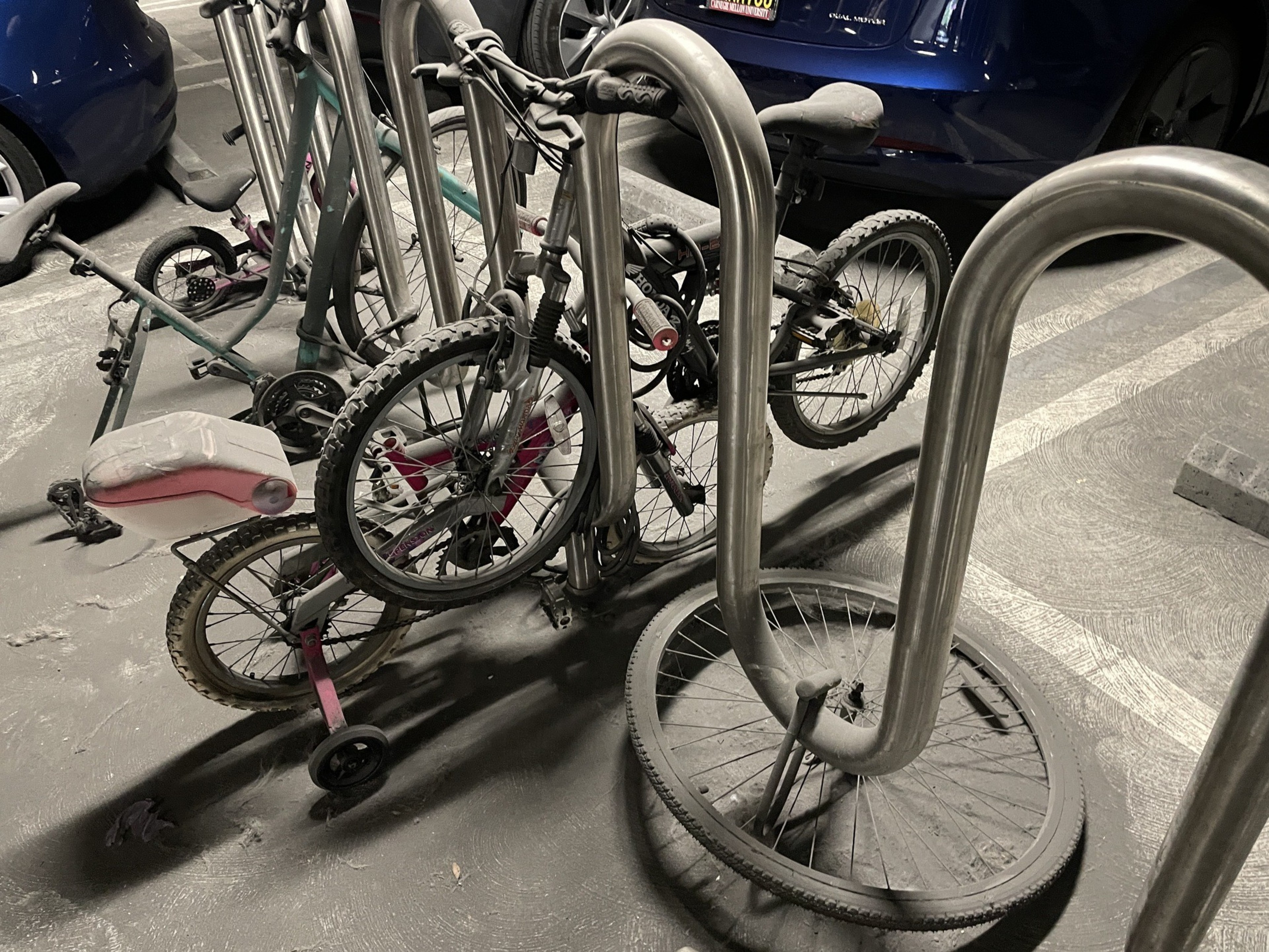 Two small bicycles, one with training wheels and a pink seat, are locked and tangled at a metal bike rack in a parking garage, with a loose front wheel nearby.