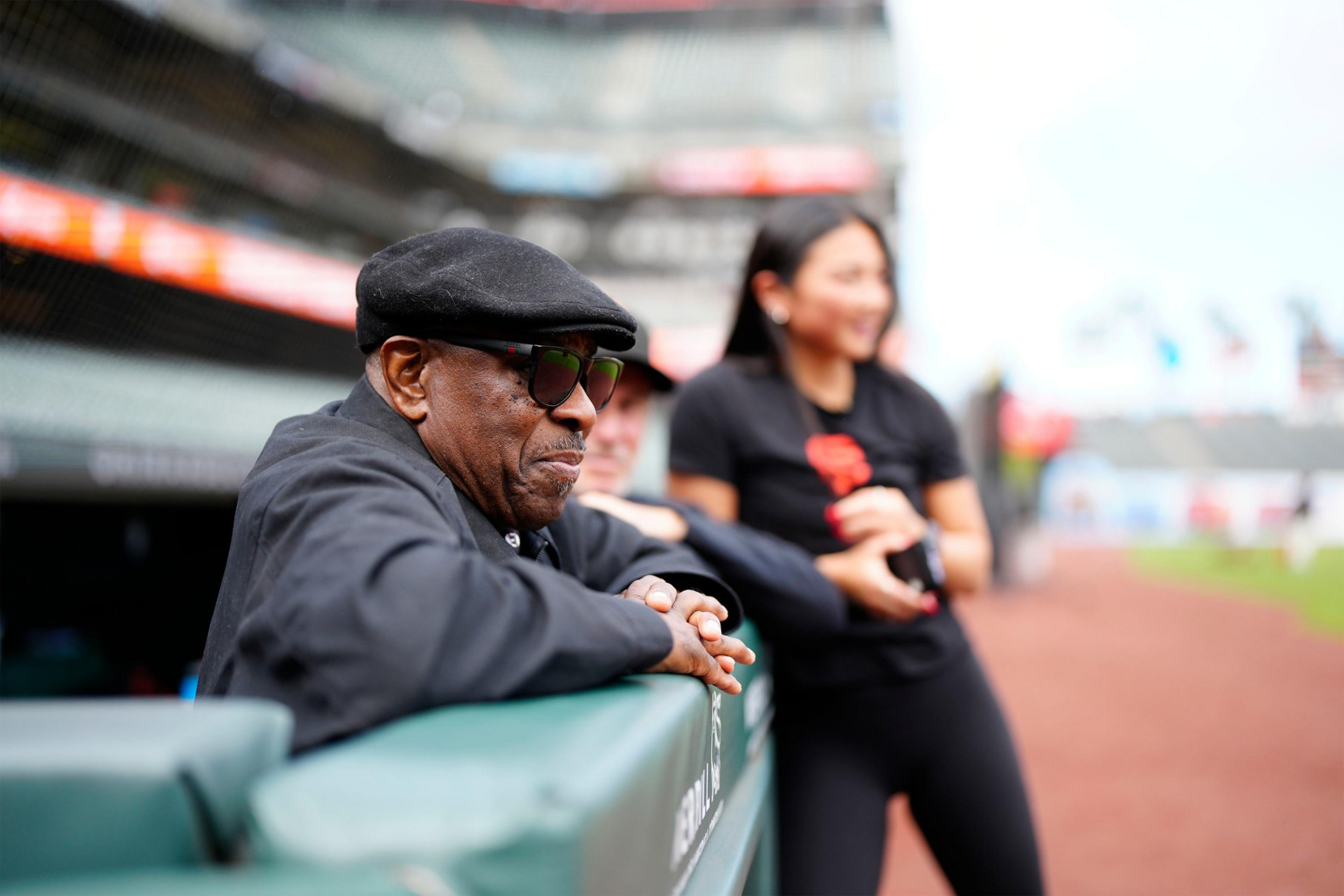 An older man wearing a black cap and sunglasses leans on a green dugout railing while a woman in black, holding a phone, stands blurred behind him.