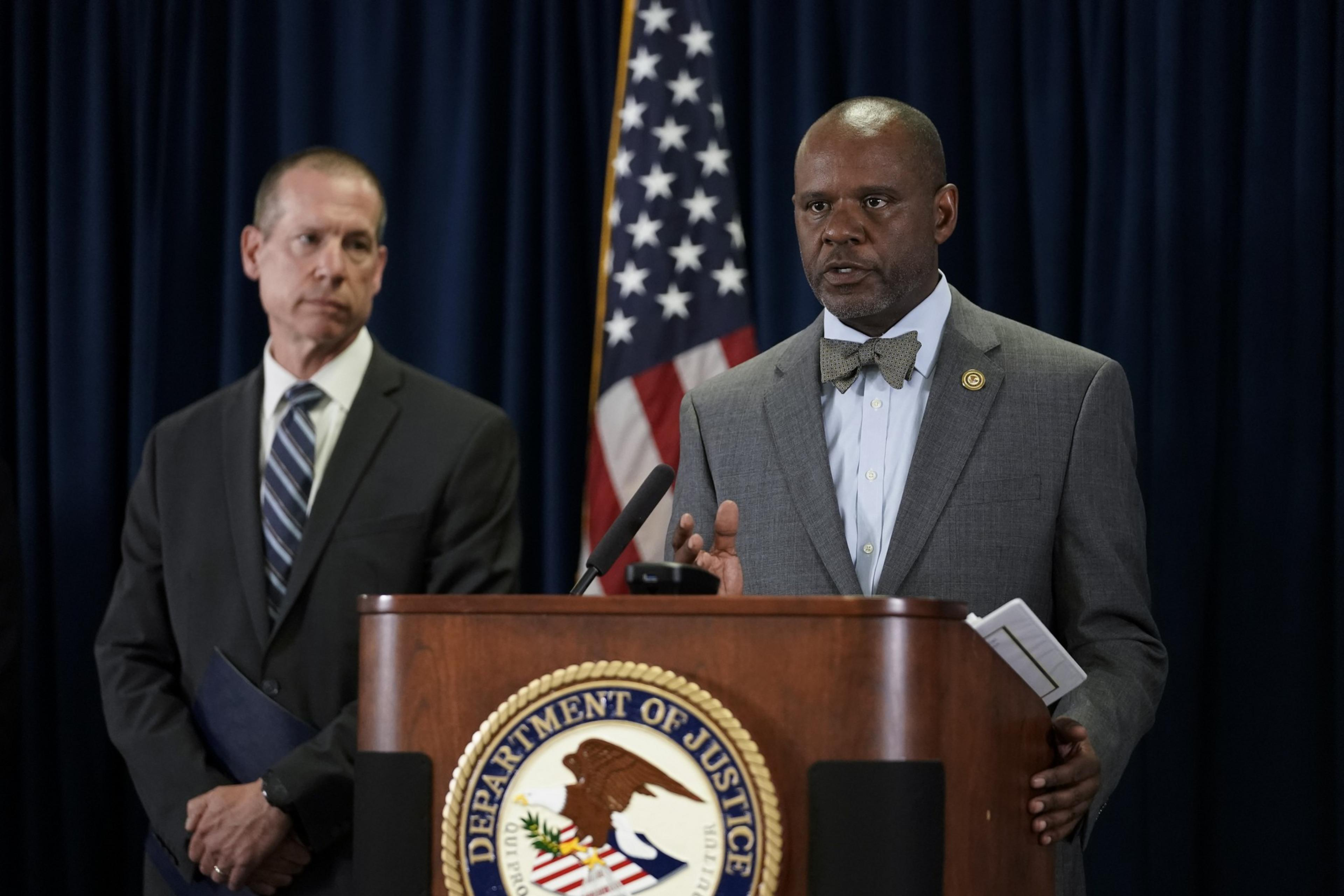 Two men in suits stand at a podium with the Department of Justice emblem. The man on the right speaks, while the one on the left listens. An American flag is behind them.