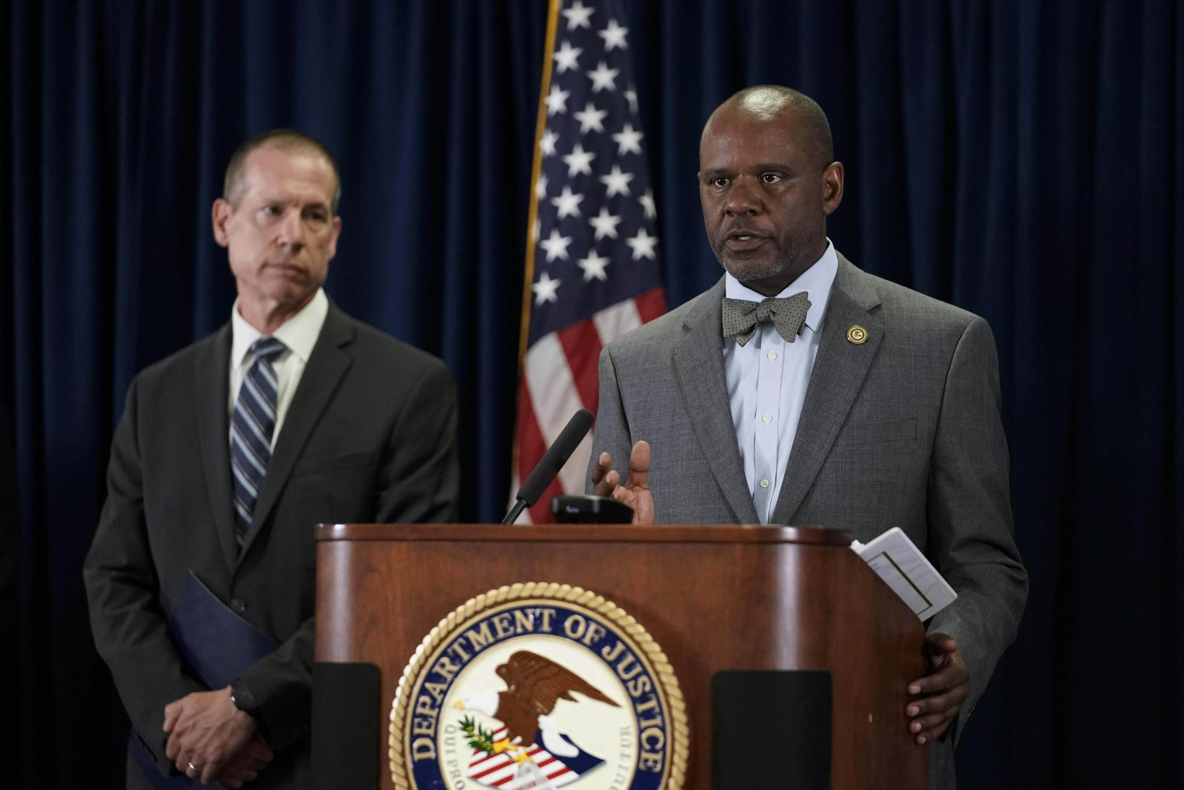 Two men in suits stand at a podium with the Department of Justice emblem. The man on the right speaks, while the one on the left listens. An American flag is behind them.