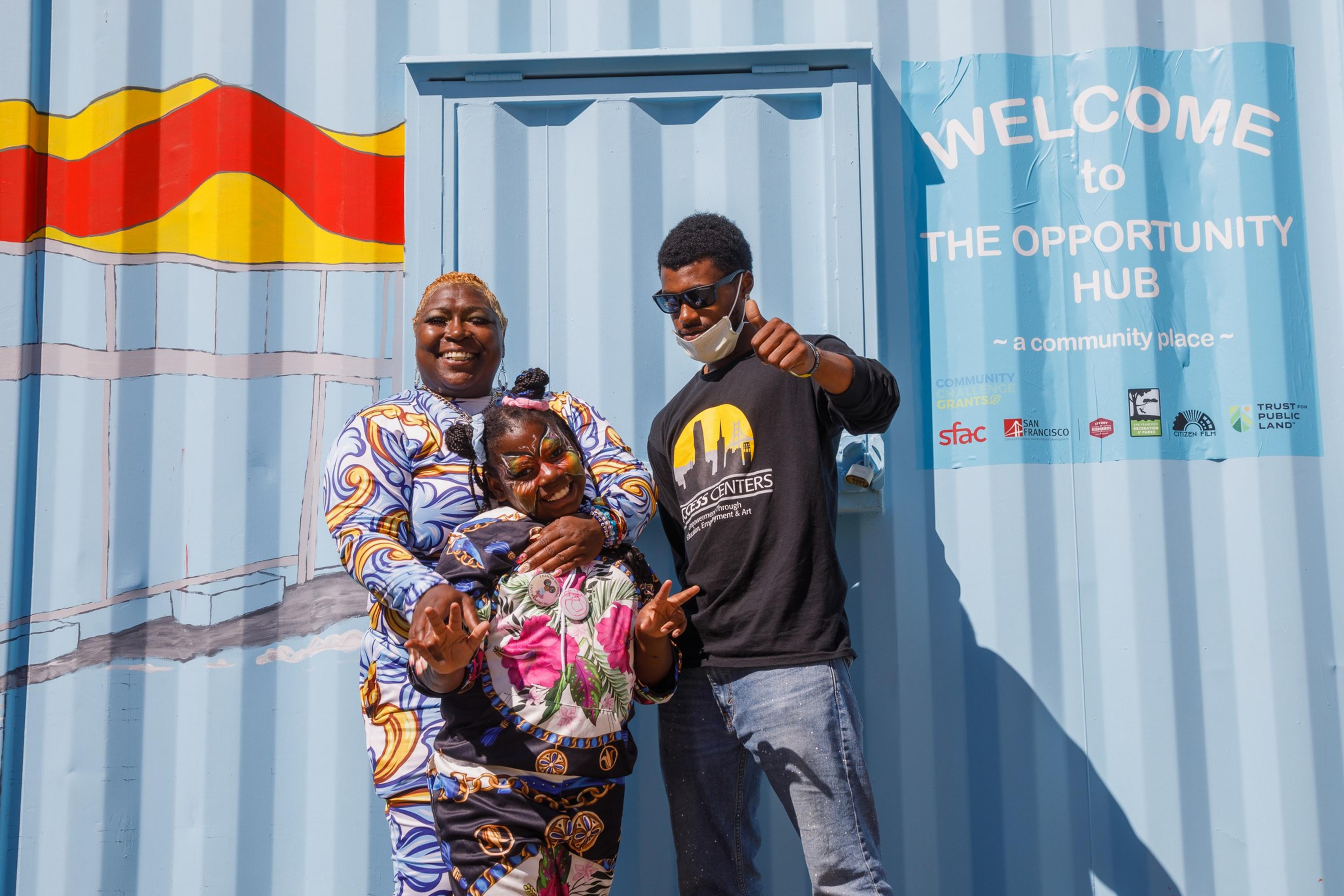 Three people are joyfully posing in front of a blue shipping container with a colorful mural and a welcoming sign for an opportunity hub.