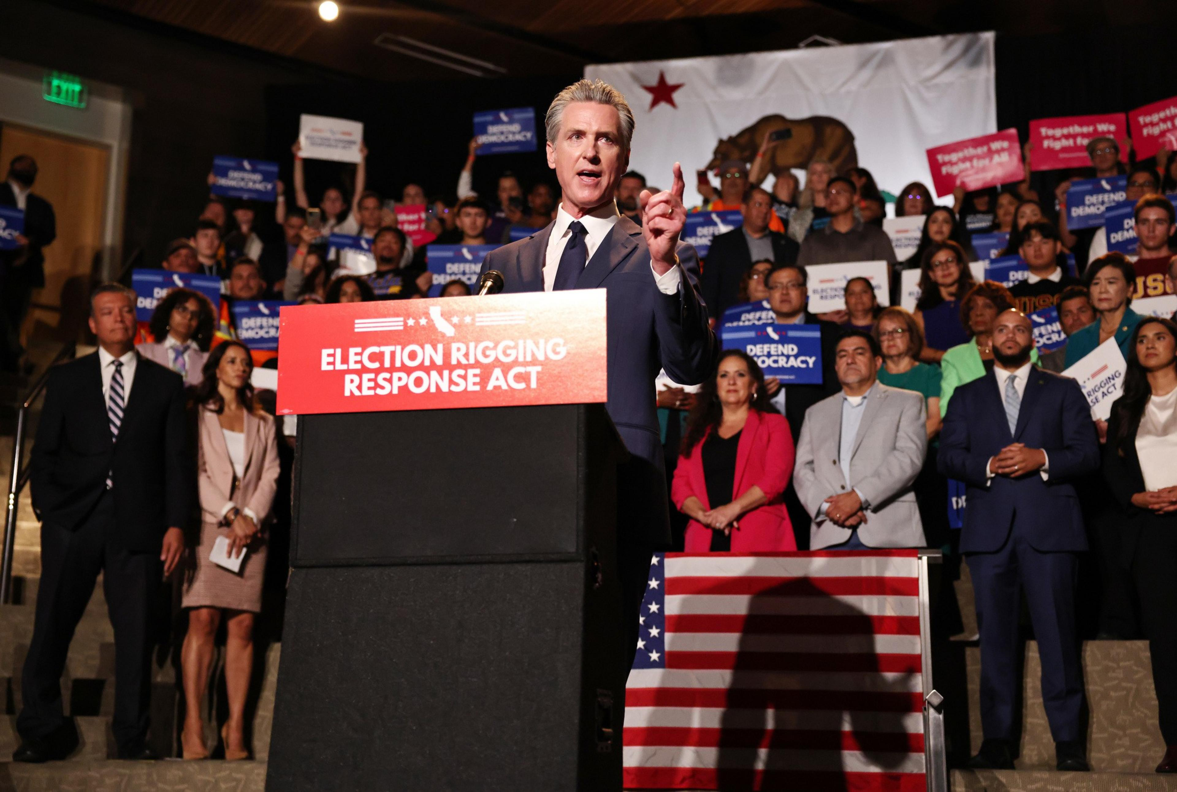 A man in a suit speaks at a podium labeled “Election Rigging Response Act,” with a crowd behind him holding signs supporting democracy and voting rights.