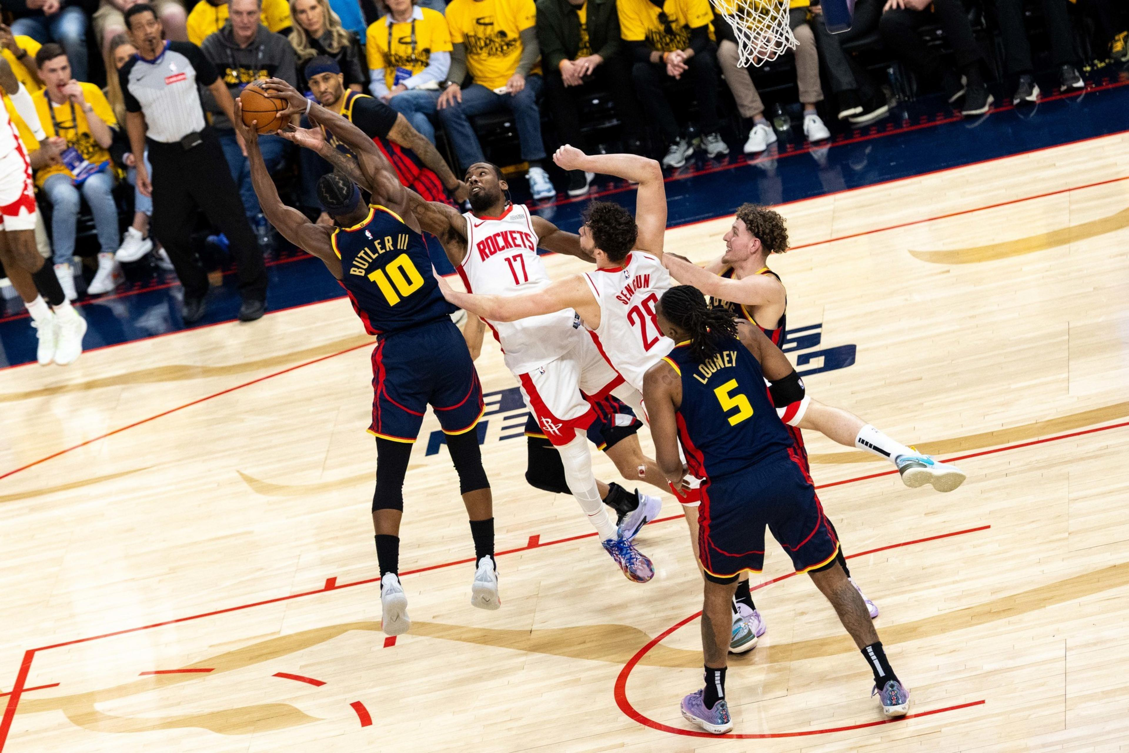 Basketball players from two teams are jumping for the ball on a court, with players in blue and yellow defending against those in white and red. Spectators watch intently.