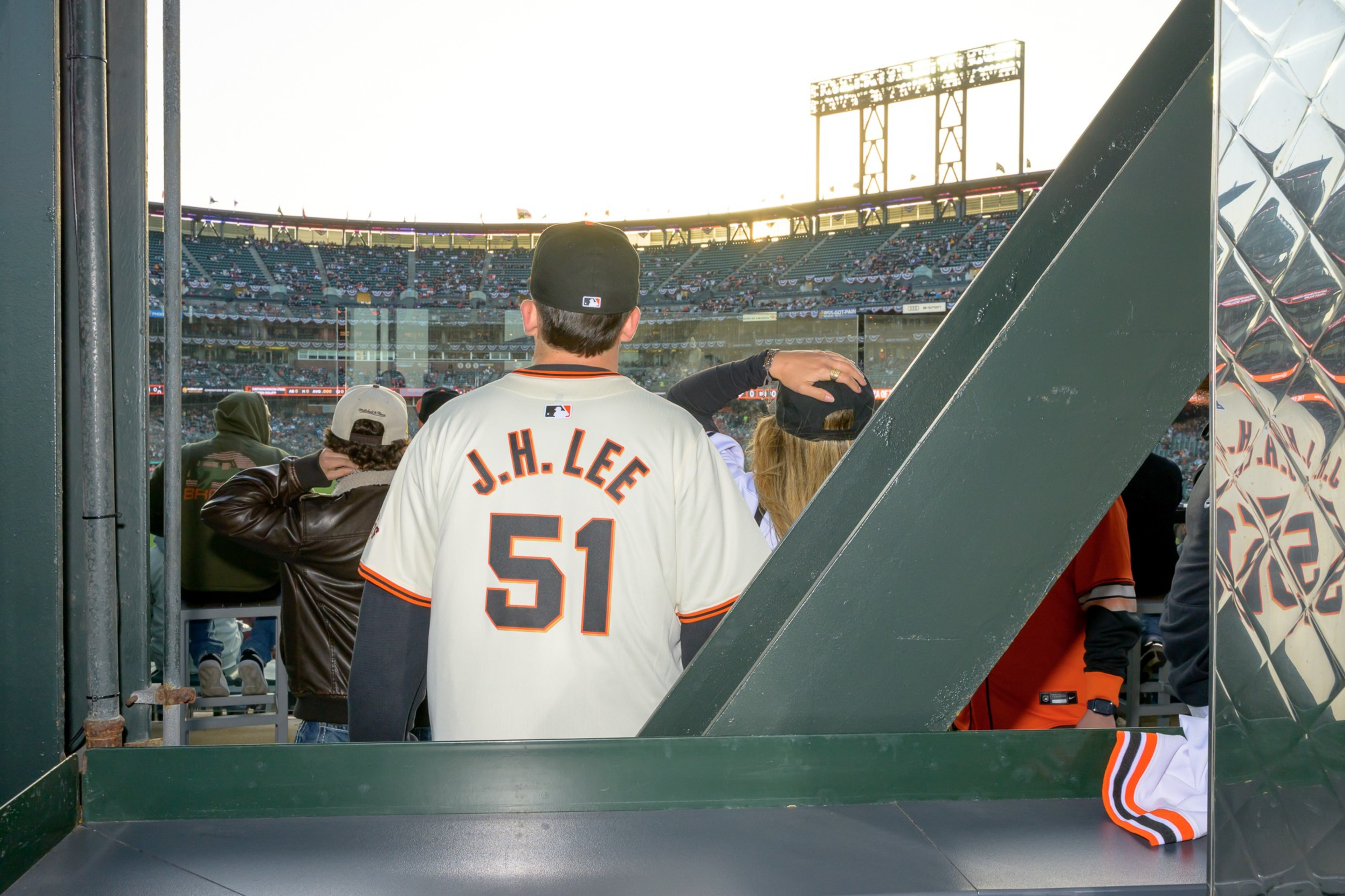 A baseball fan wearing a jersey labeled &quot;J.H. Lee 51&quot; stands in a stadium viewing a game, surrounded by other spectators and structural elements.
