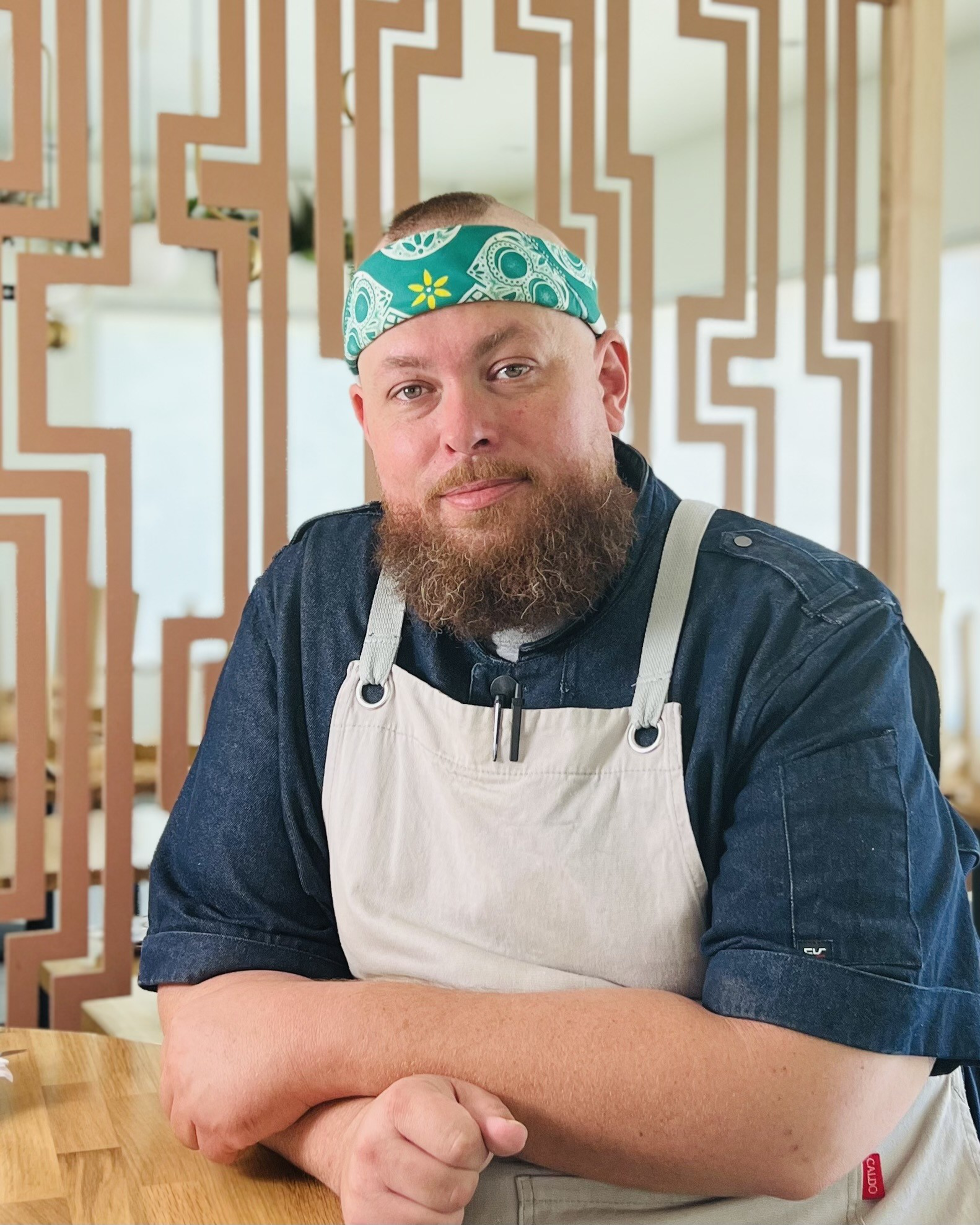 A bearded man wearing a teal patterned bandana and dark shirt with a beige apron sits at a wooden table, arms crossed, looking calmly at the camera.