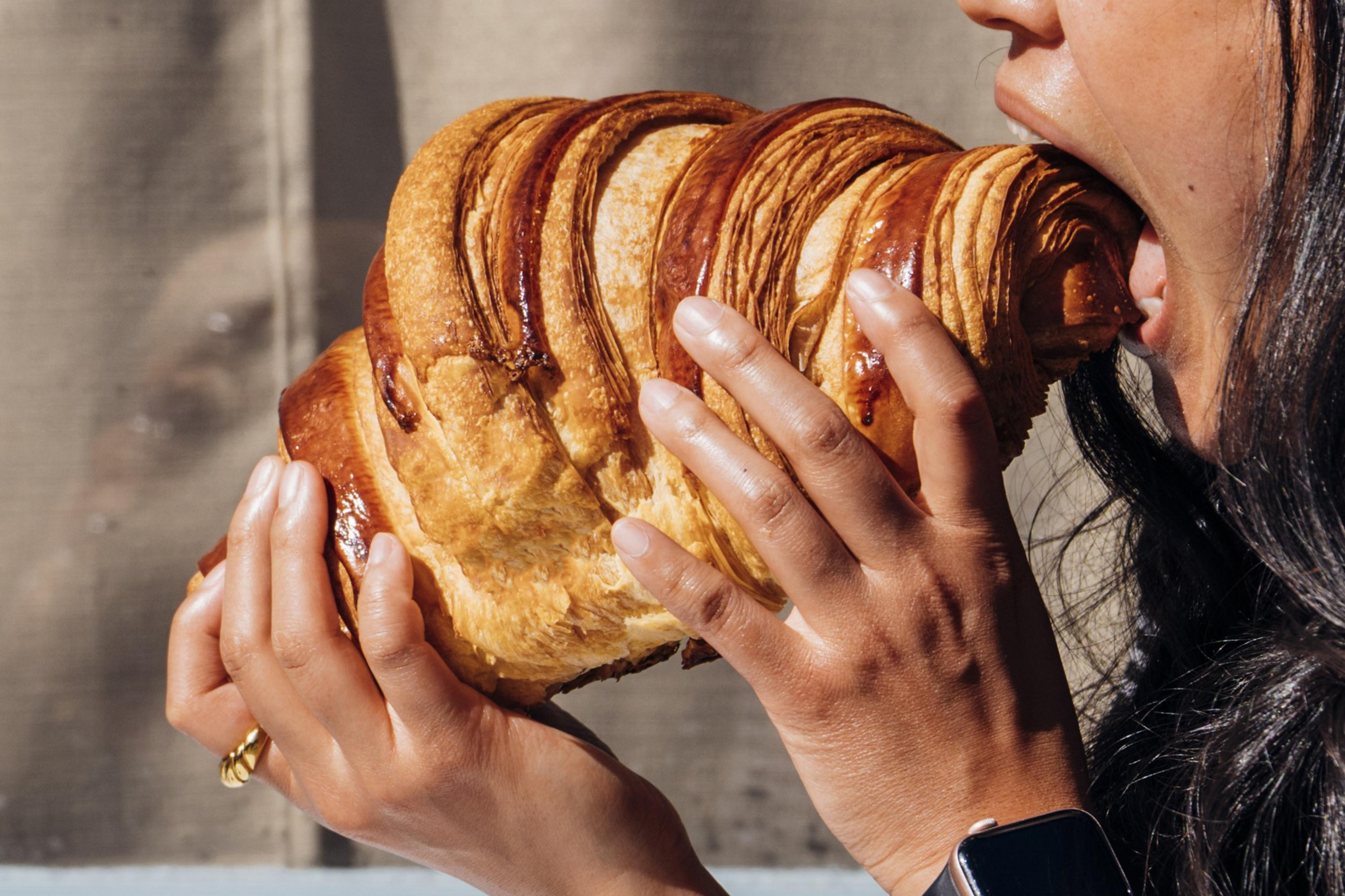 A person is about to bite into an oversized croissant, holding it with both hands. Their hair is dark, and they have a smartwatch on their wrist.