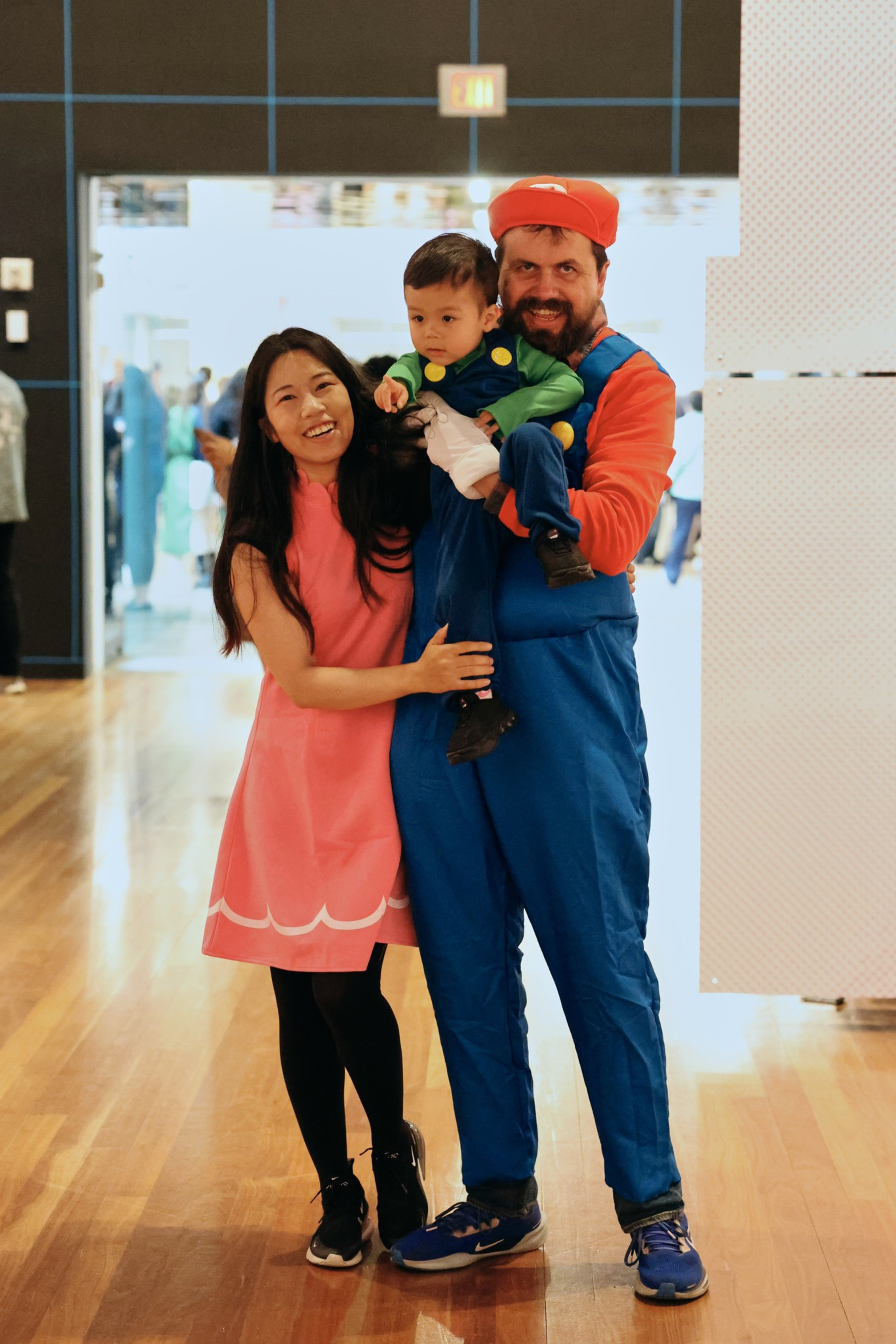 A man dressed as Mario holds a toddler dressed as Luigi, while a woman in a pink dress stands smiling beside them on a wooden floor.