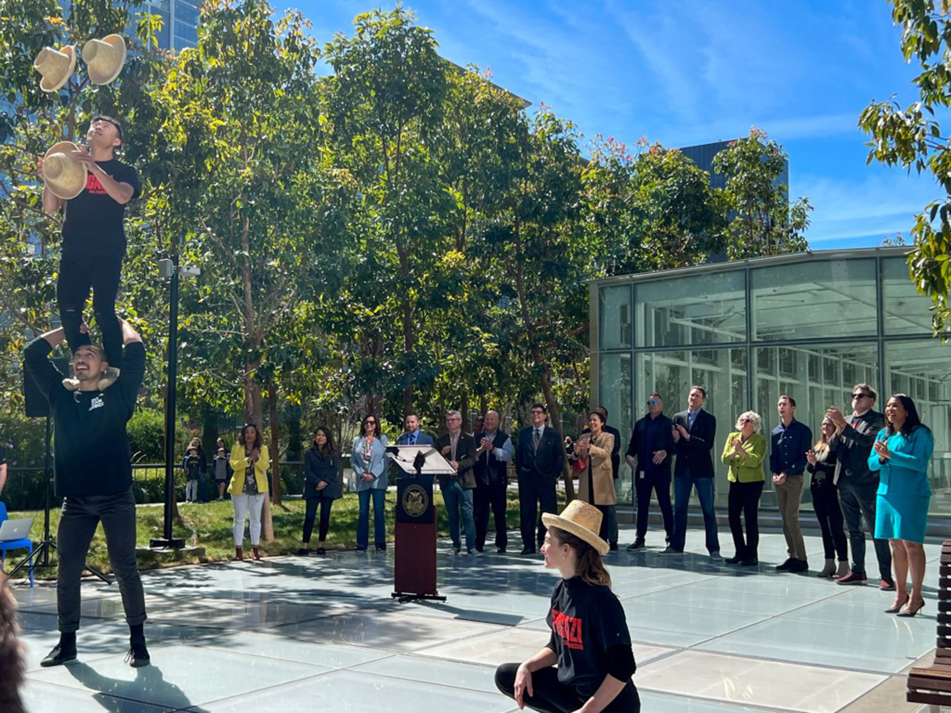 Two performers stack hats while one stands on another's shoulders, as a crowd of people in formal and casual attire watches and applauds outdoors near trees and a glass building.