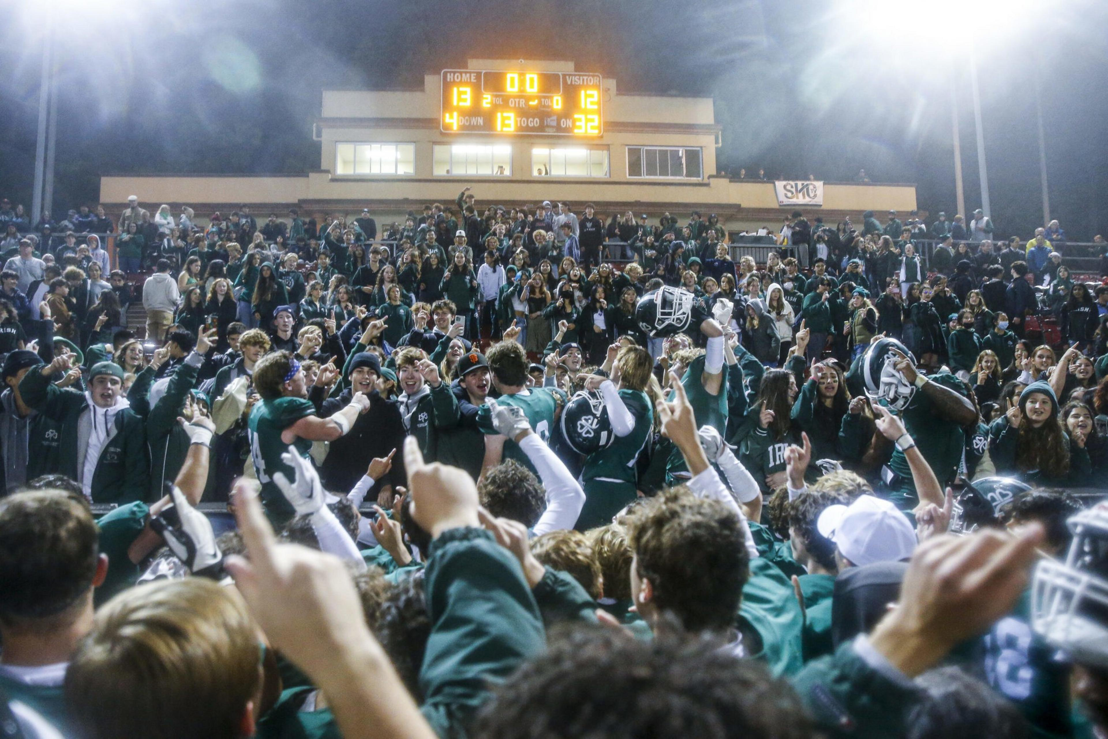 A football team and fans celebrate a narrow 13-12 victory under bright stadium lights, with many raising their hands in excitement.