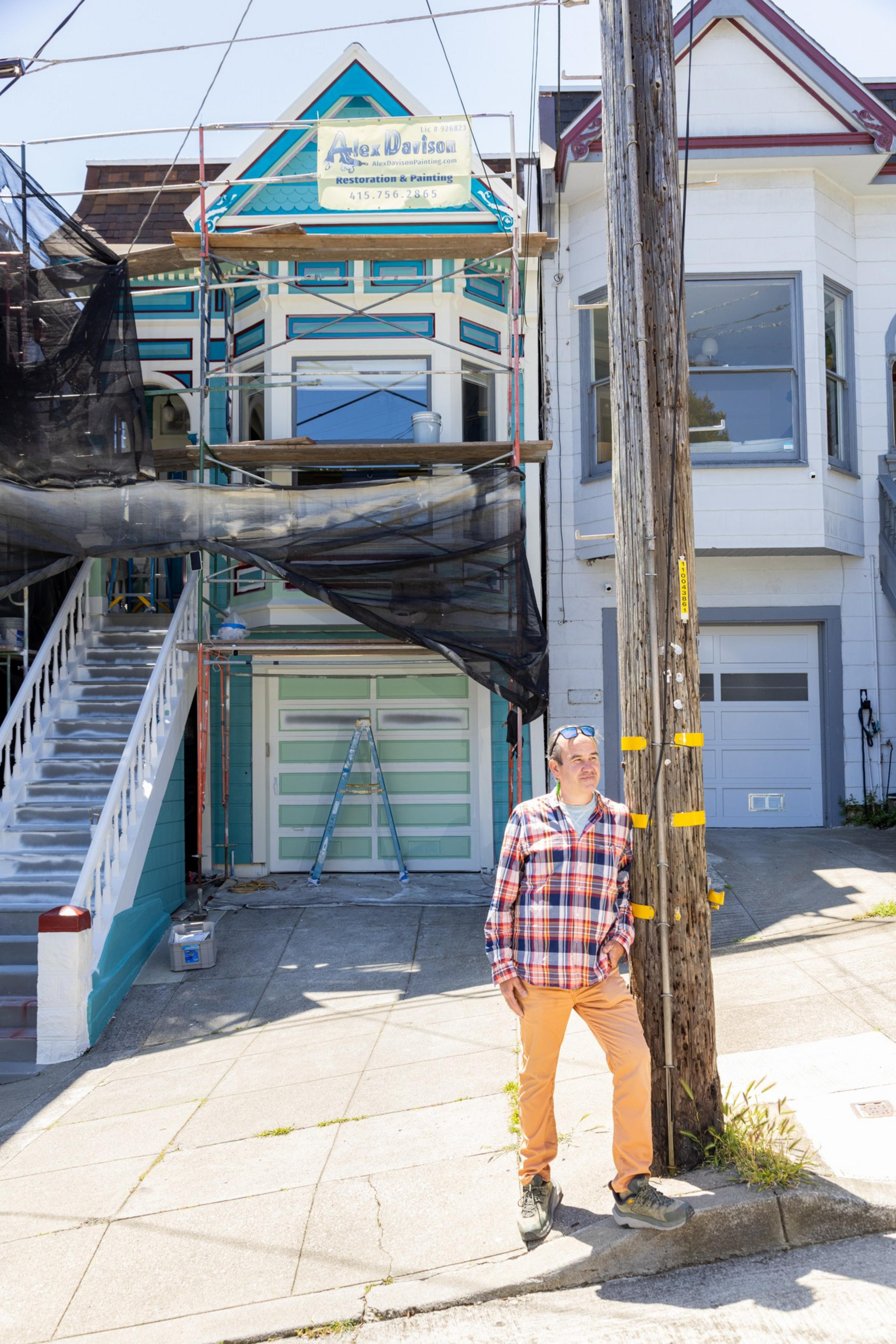 A man stands before a colorful, partly scaffolded Victorian house with a clear blue sky behind.