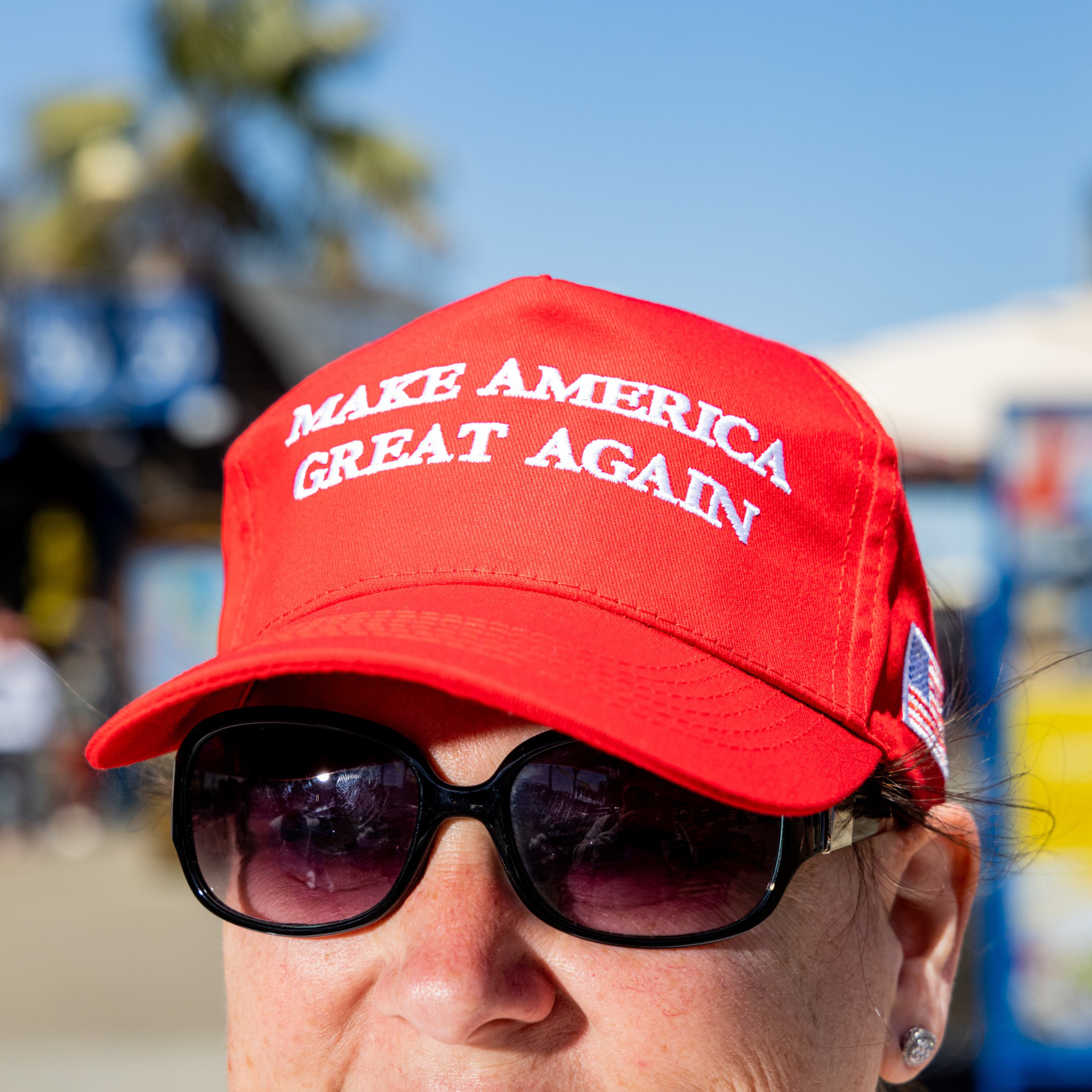 A person wearing a bright red cap with the slogan &quot;MAKE AMERICA GREAT AGAIN&quot; in white letters, sunglasses, and an American flag patch on the side.