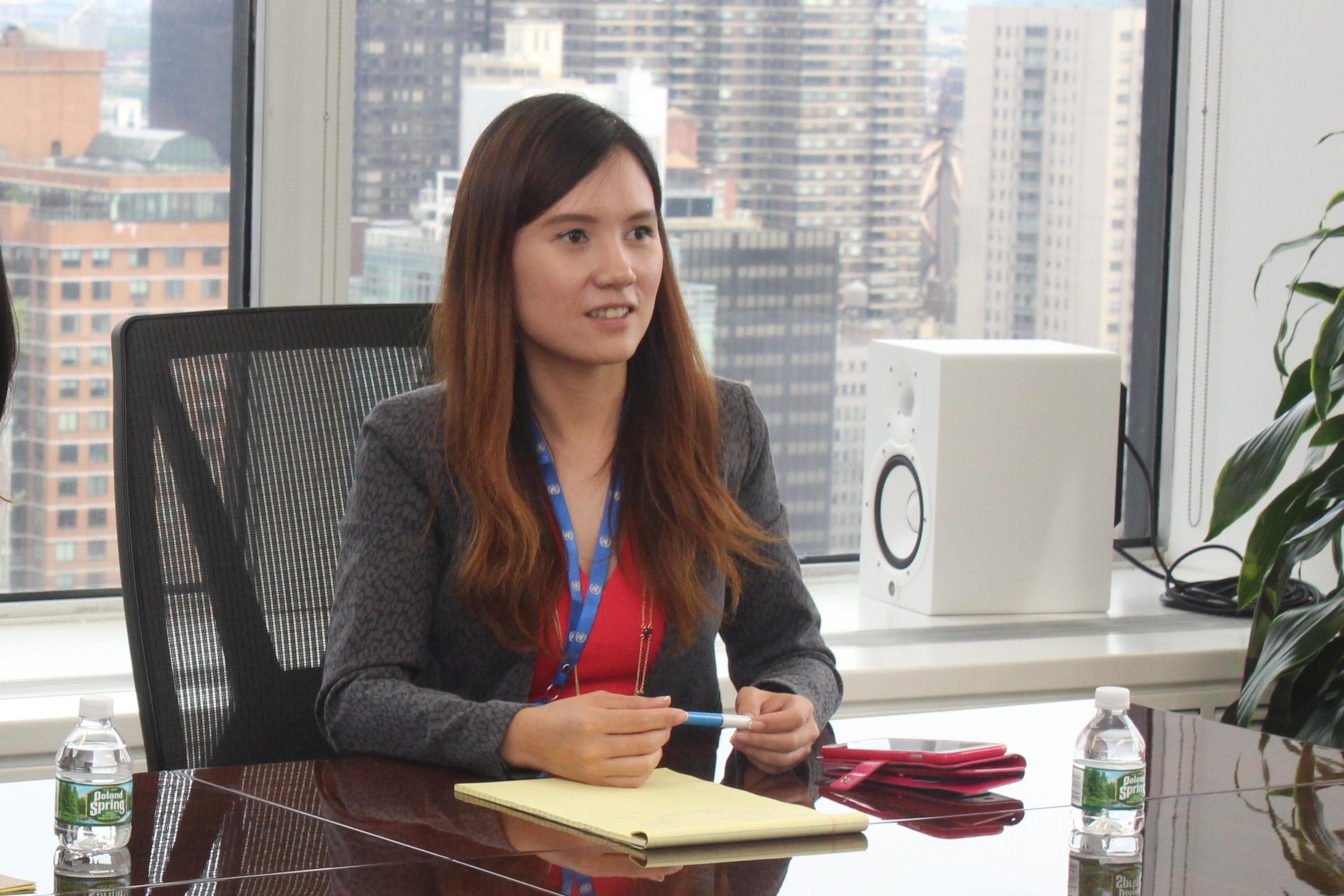 A woman with long brown hair wearing a gray blazer and red shirt sits at a conference table holding a pen, with a cityscape visible through large windows.