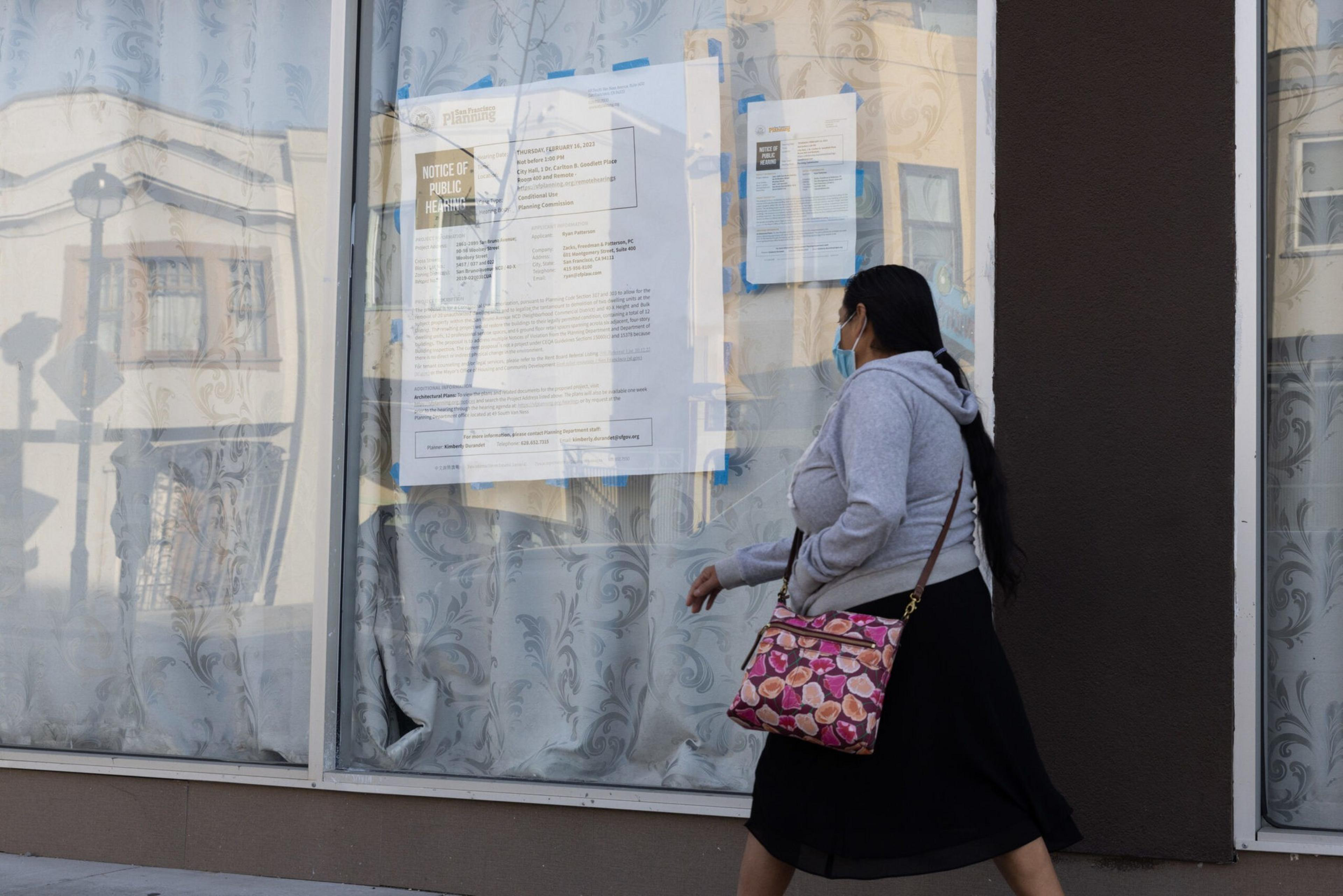 A woman with long black hair, wearing a mask, gray hoodie, and black skirt, walks past a window covered with patterned curtains and posted notices.