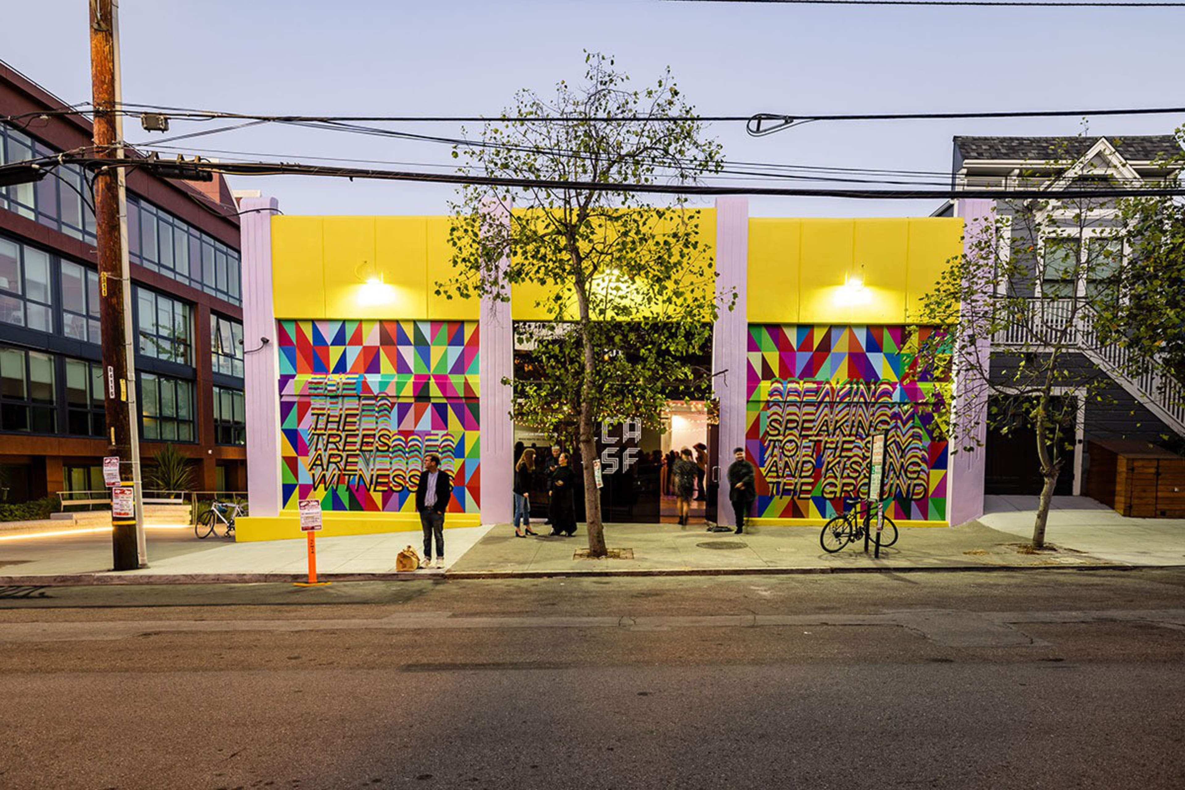 A yellow building with colorful geometric murals and text, a central tree, people gathered inside, and a bike parked on the sidewalk.