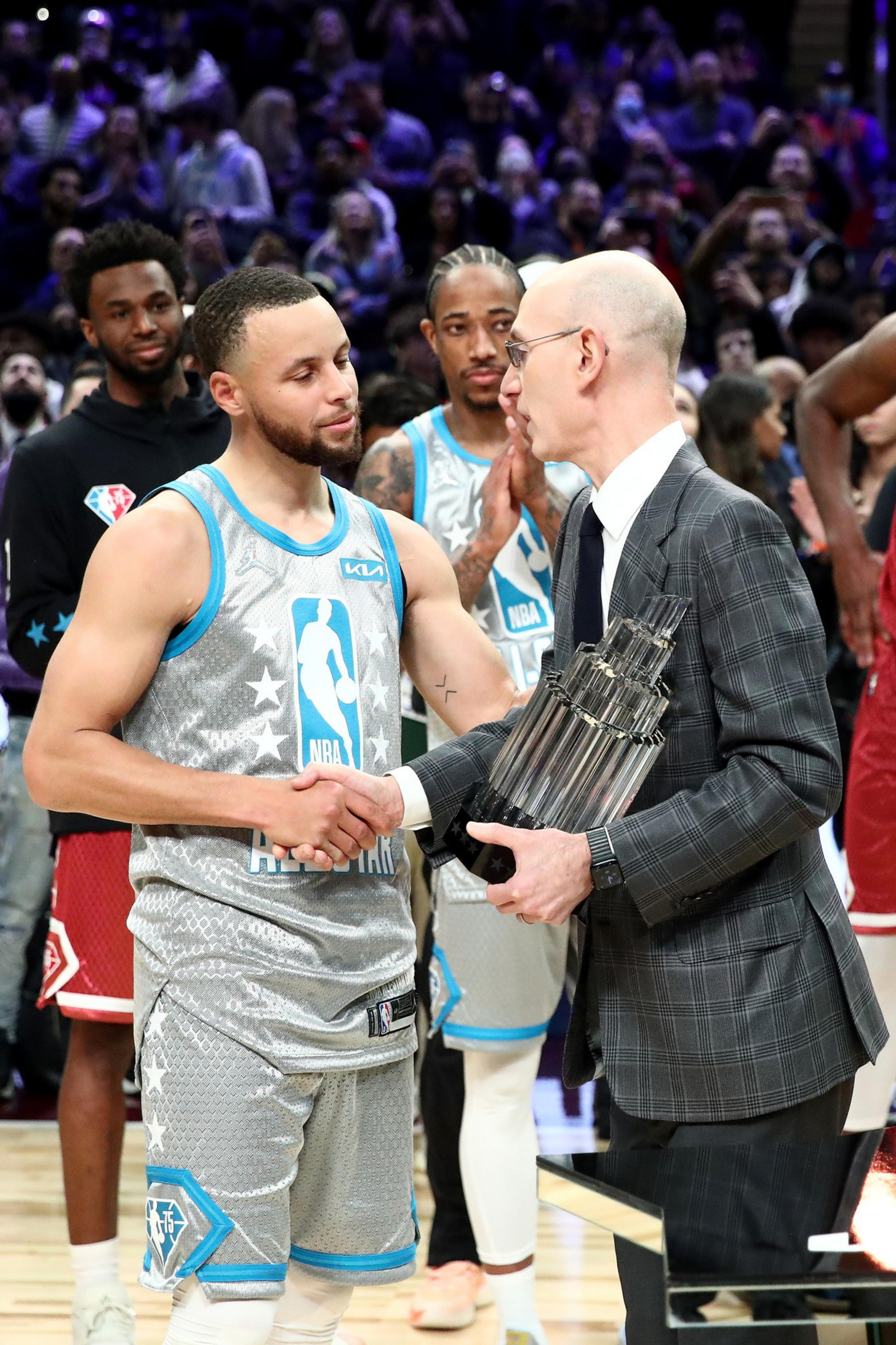 A basketball player in a gray All-Star jersey shakes hands with a man in a suit, who holds a trophy. Players and a crowd are seen in the background.
