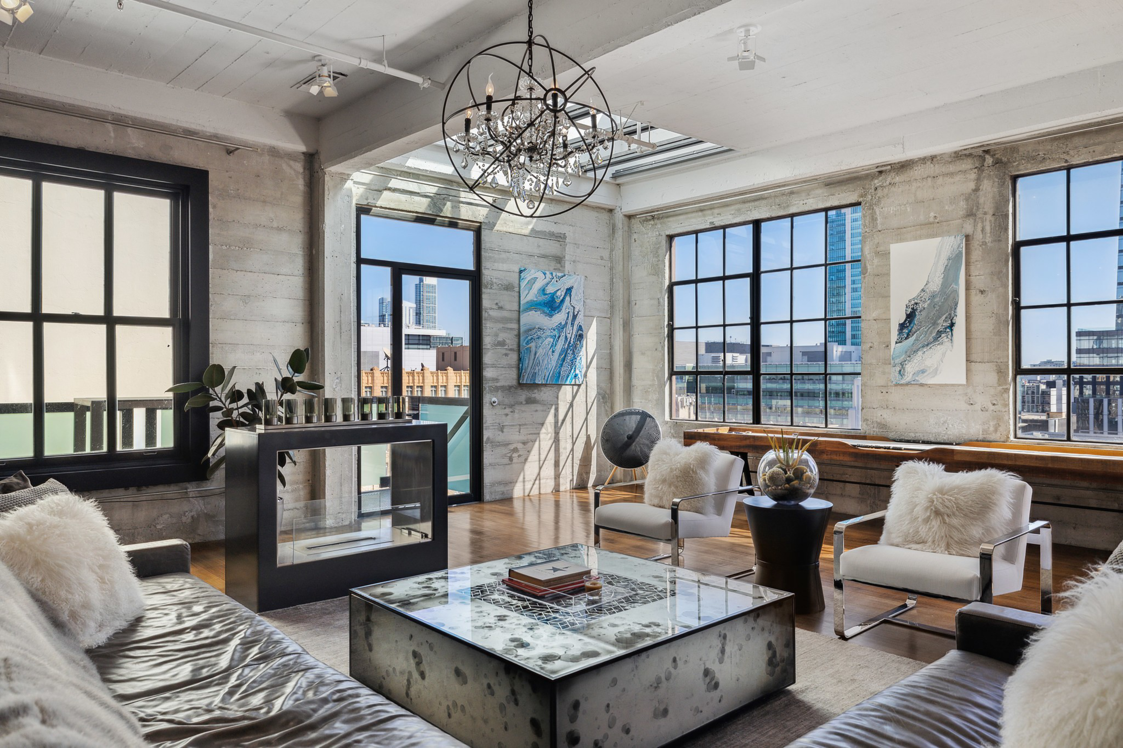A modern living room with large industrial windows, a glass skylight, a sleek metal and glass coffee table, plush white cushions, and abstract wall art.