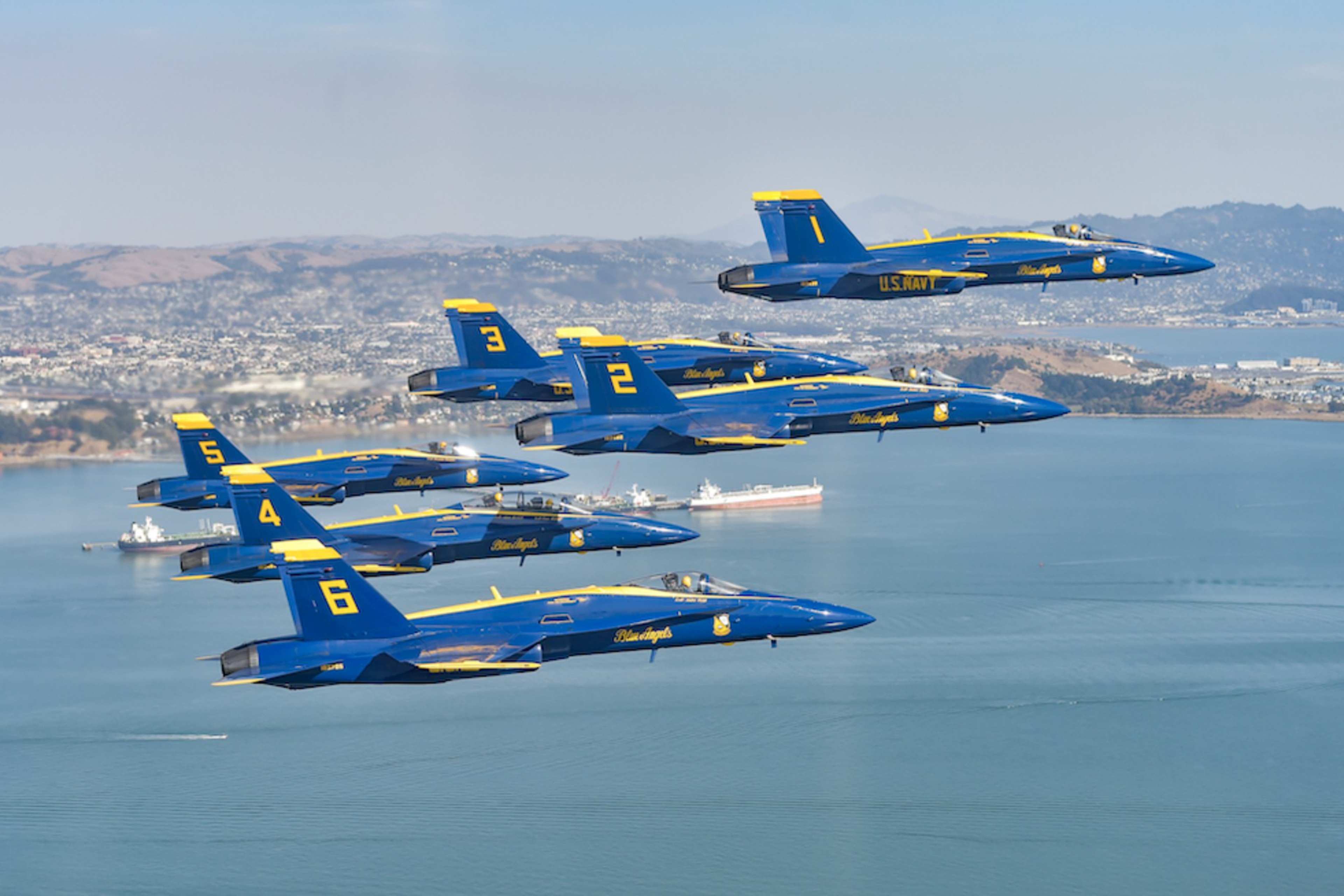 Six blue and yellow US Navy Blue Angels jets fly in tight formation over a coastal area with ships and hills in the background.