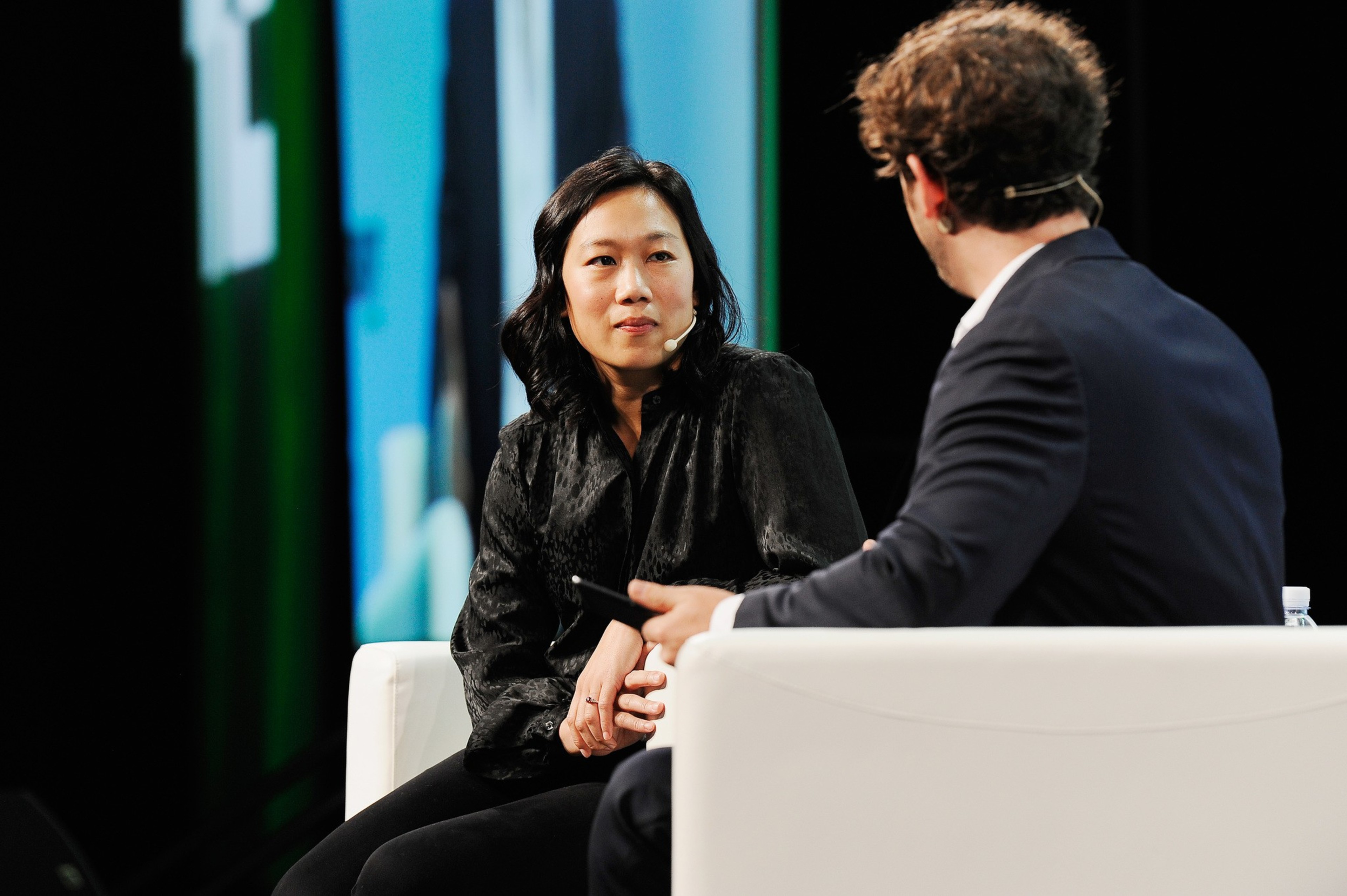 Two people are seated on a stage, engaged in conversation. Both wear microphones. The woman has long dark hair and is facing the man, who has his back to the camera.