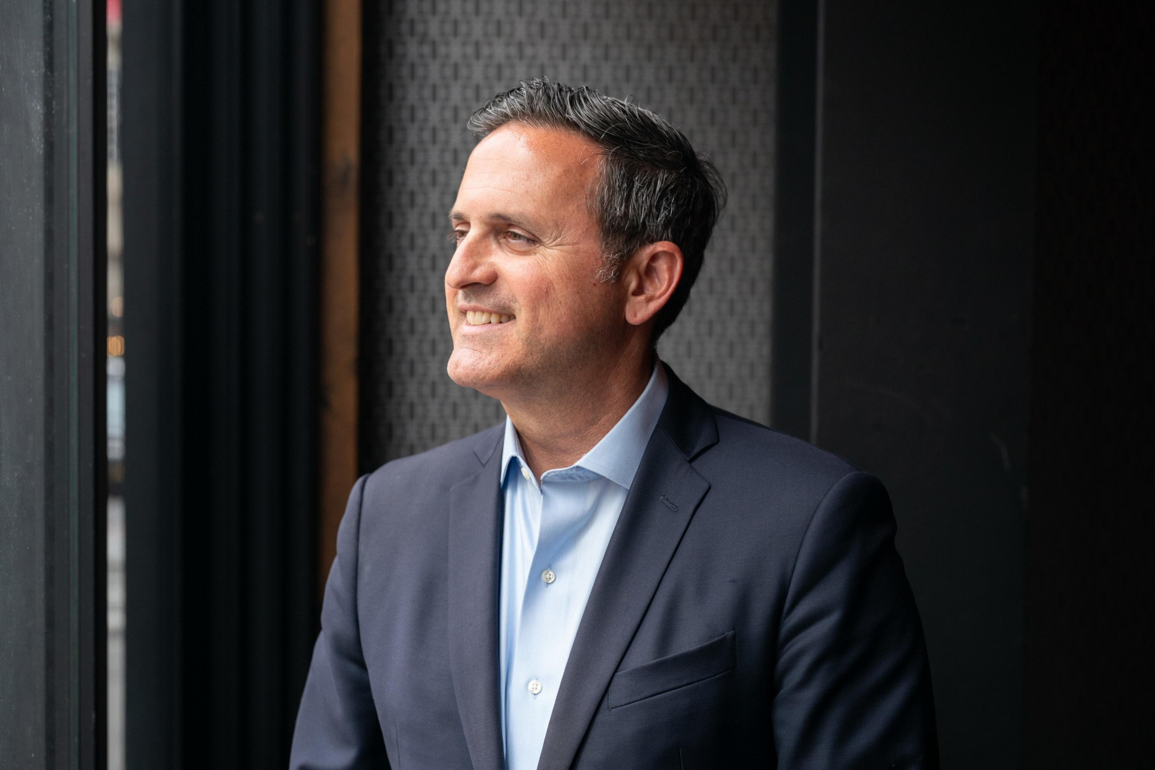 A man in a business suit smiles broadly, looking off to the side; background suggests an indoor setting with subtle decor.