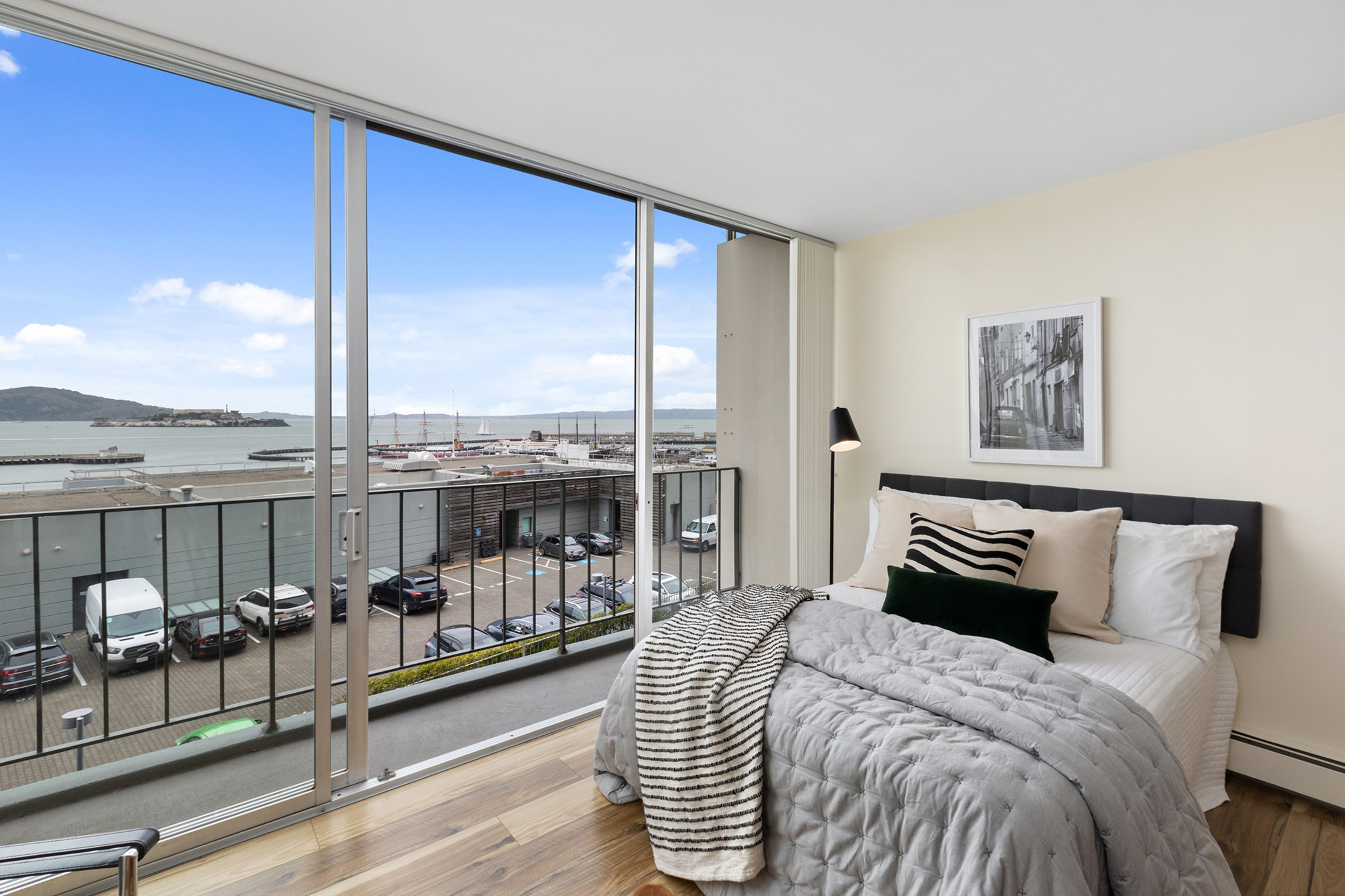 A cozy bedroom with a neatly made bed featuring gray and striped bedding, a black floor lamp, and a large window overlooking a marina and parked cars.
